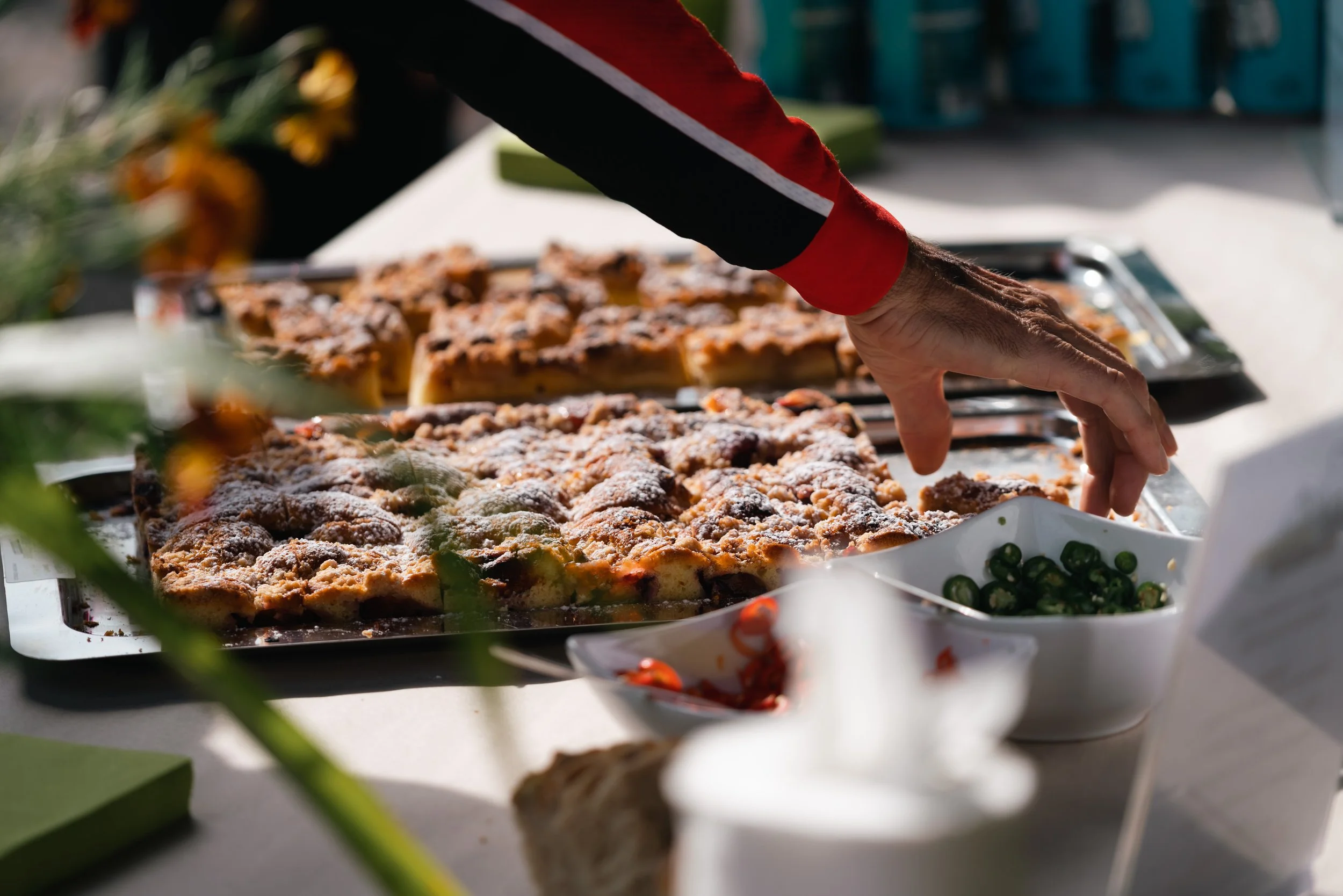 Nahaufnahme von jemandem, der mit der Hand ein Stück Streuselkuchen von einem Blech nimmt, daneben eine Schale mit geschnittenen grünen Chilis auf einem Tisch.