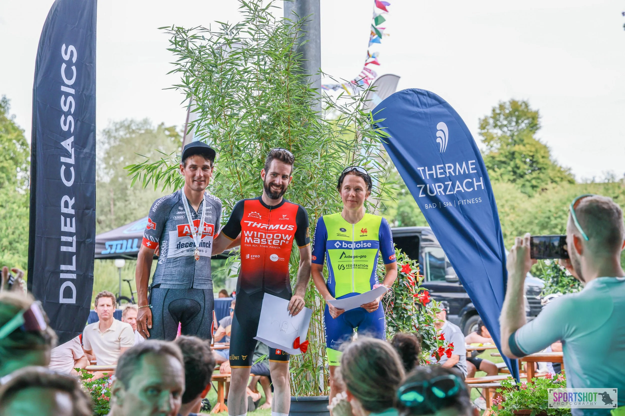 Drei Radfahrer in Sportkleidung stehen auf einem Podium bei einer Siegerehrung. Zwei Werbeflaggen im Hintergrund, eine mit der Aufschrift 'Therme Zurzach'. Zuschauer sitzen und stehen in der Nähe. Bunte Wimpel hängen über dem Podium.