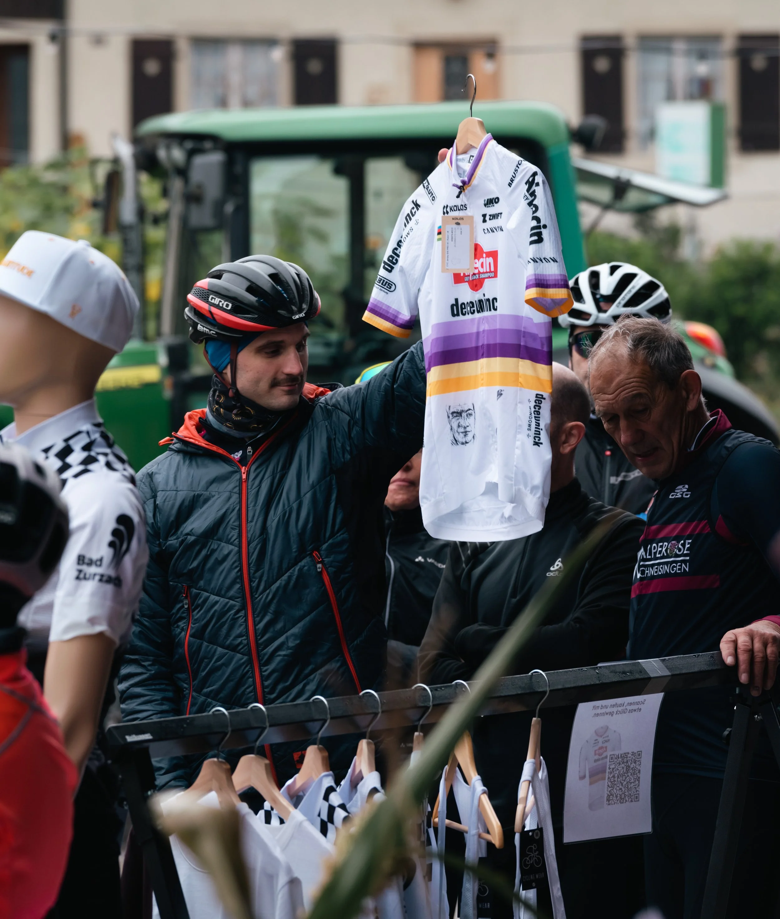 Person hält ein Radtrikot in die Höhe, umgeben von anderen Menschen und einem Mannequin mit Radbekleidung, Outdoor-Umgebung.