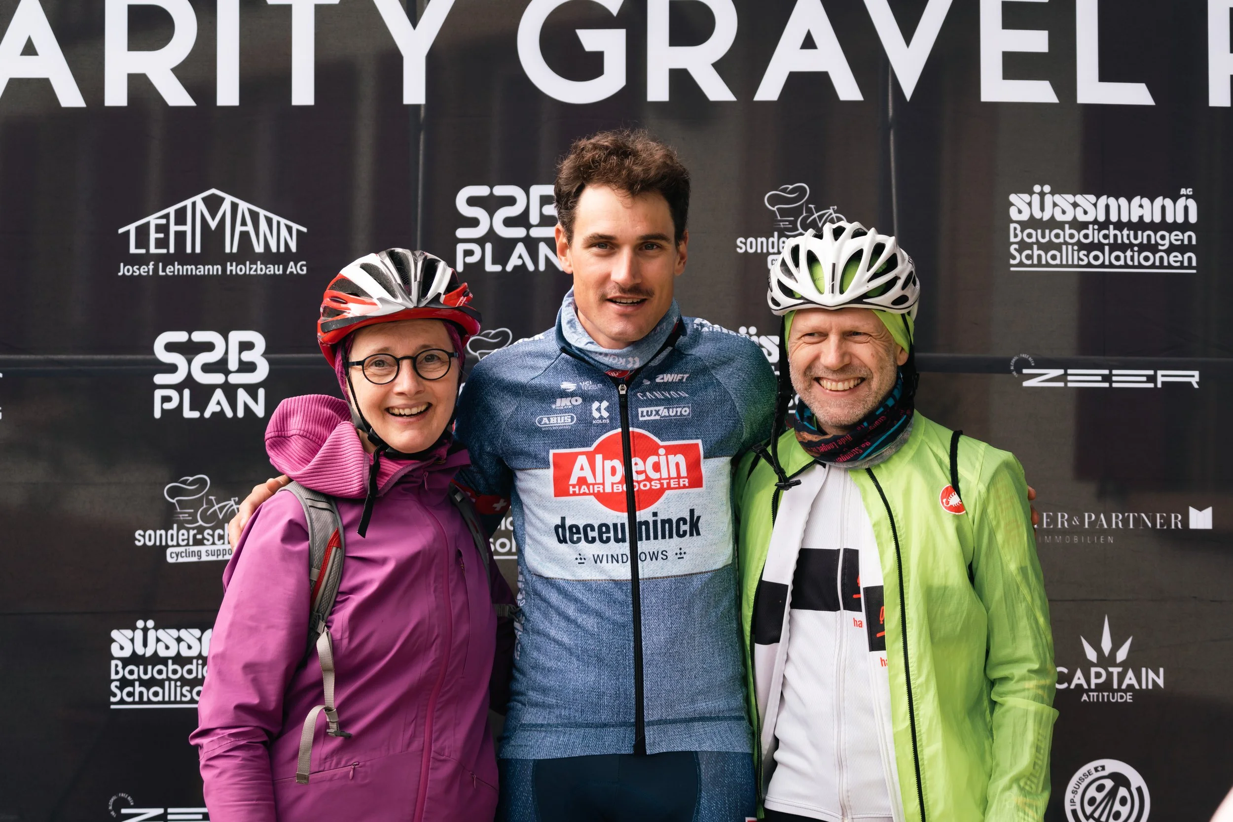 Drei Personen in Radkleidung stehen vor einem Banner mit Sponsorenlogos. Die zentral stehende Person trägt ein Alpecin Deceuninck Trikot, die anderen beiden Personen tragen Helme und Jacken in Rot und Grün.