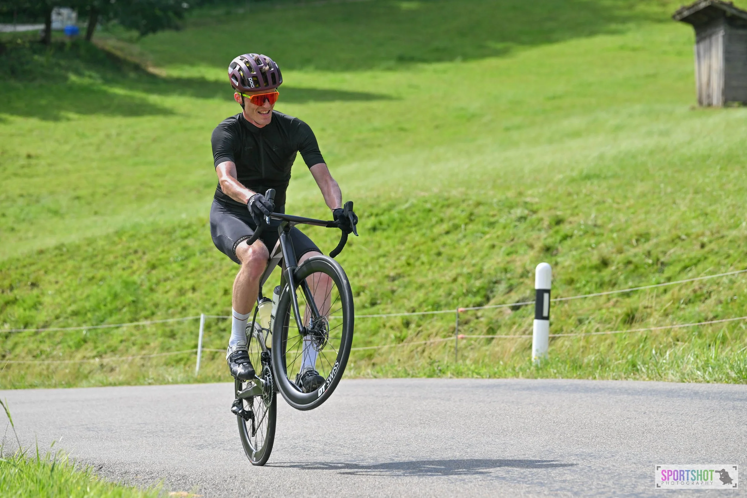 Ein Radfahrer in schwarzer Sportbekleidung und Helm macht einen Wheelie auf seinem Rennrad auf einer ländlichen Straße.
