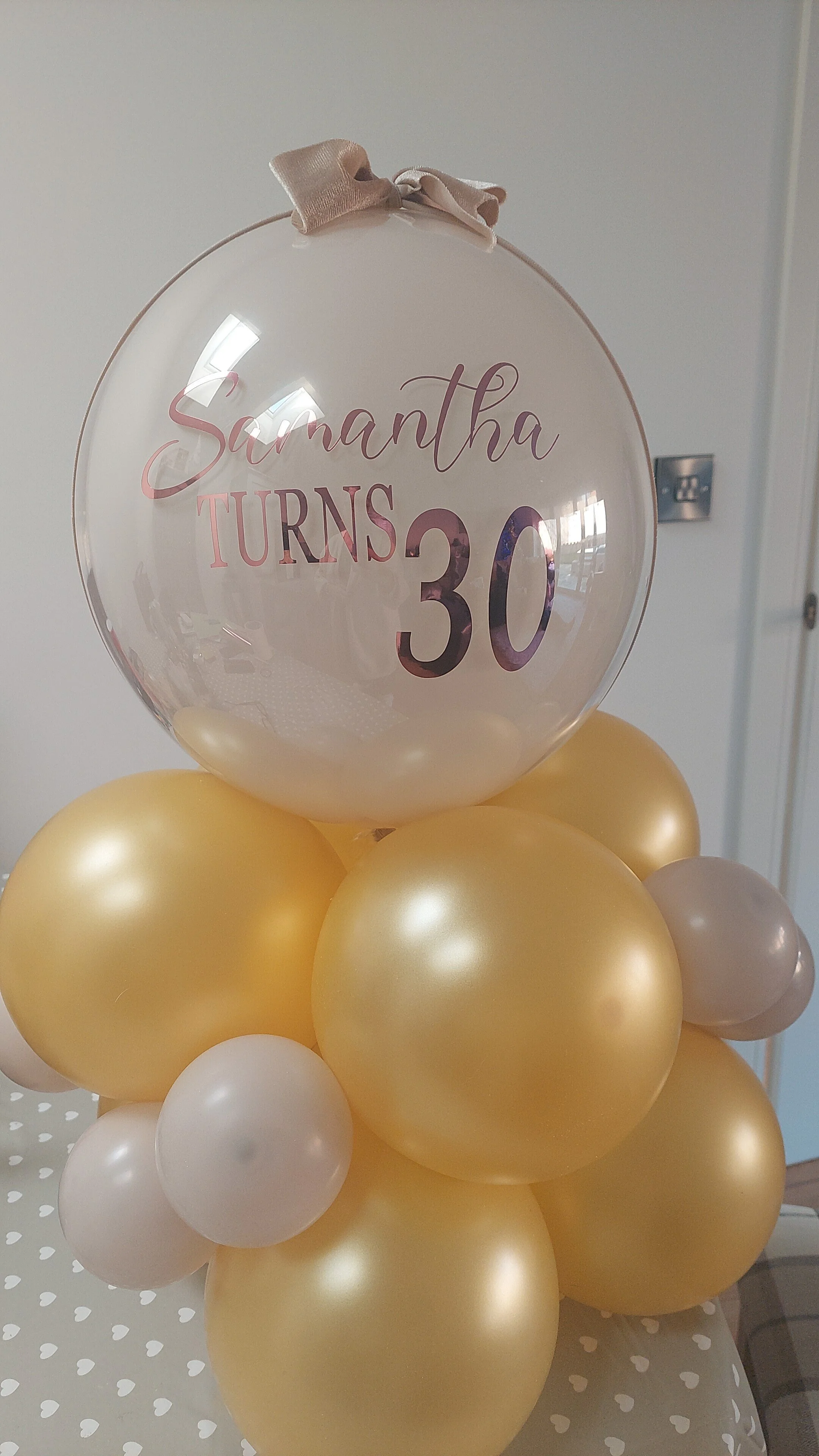 A balloon arrangement featuring a transparent balloon with the message 'Samantha Turns 30' printed inside, topped with a beige ribbon, attached to a cluster of gold and white balloons, on a table covered with a gray tablecloth with white hearts.