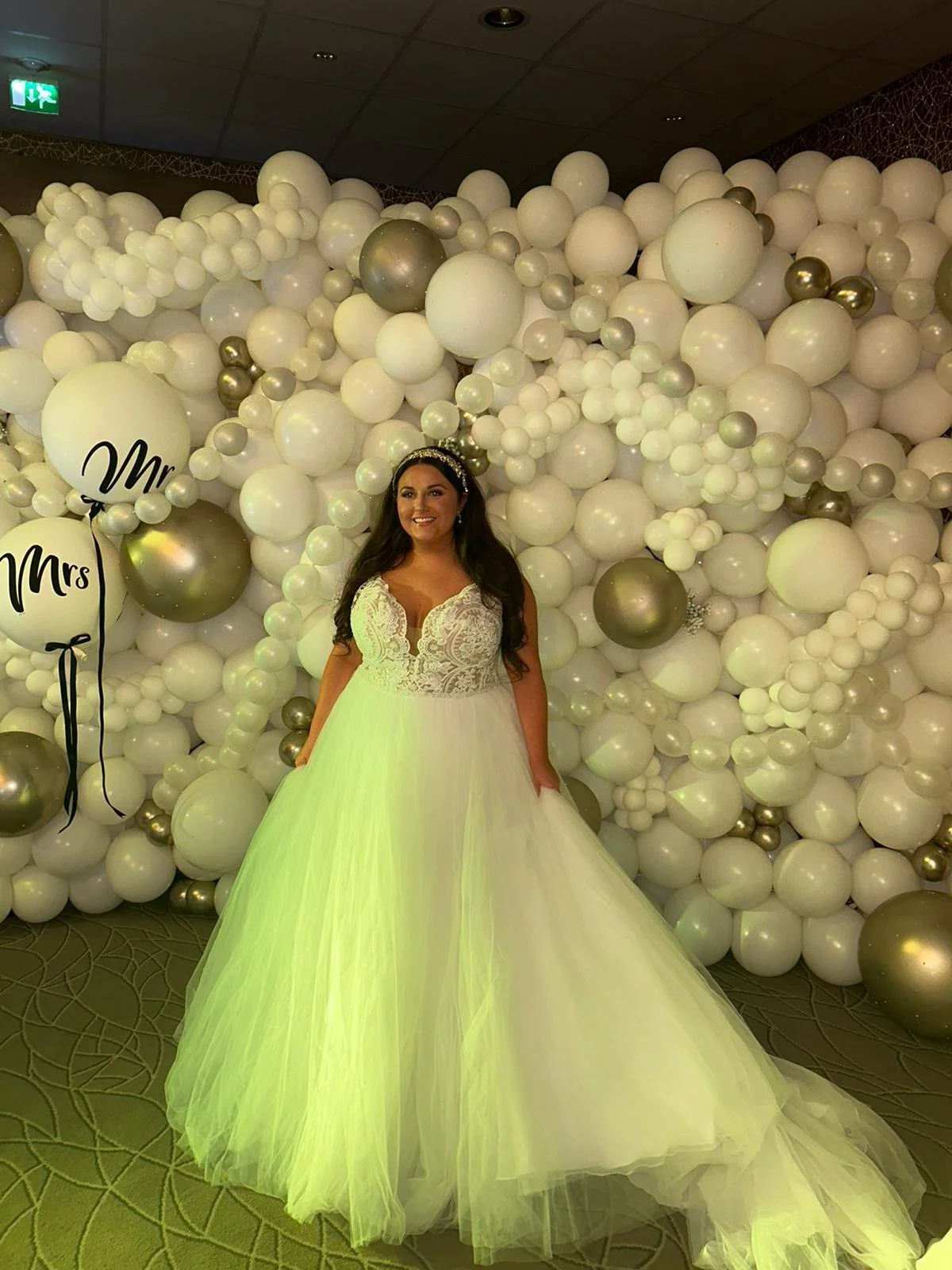 Woman in a white wedding dress standing in front of a balloon backdrop with white, gold, and silver balloons, posing at a wedding celebration.