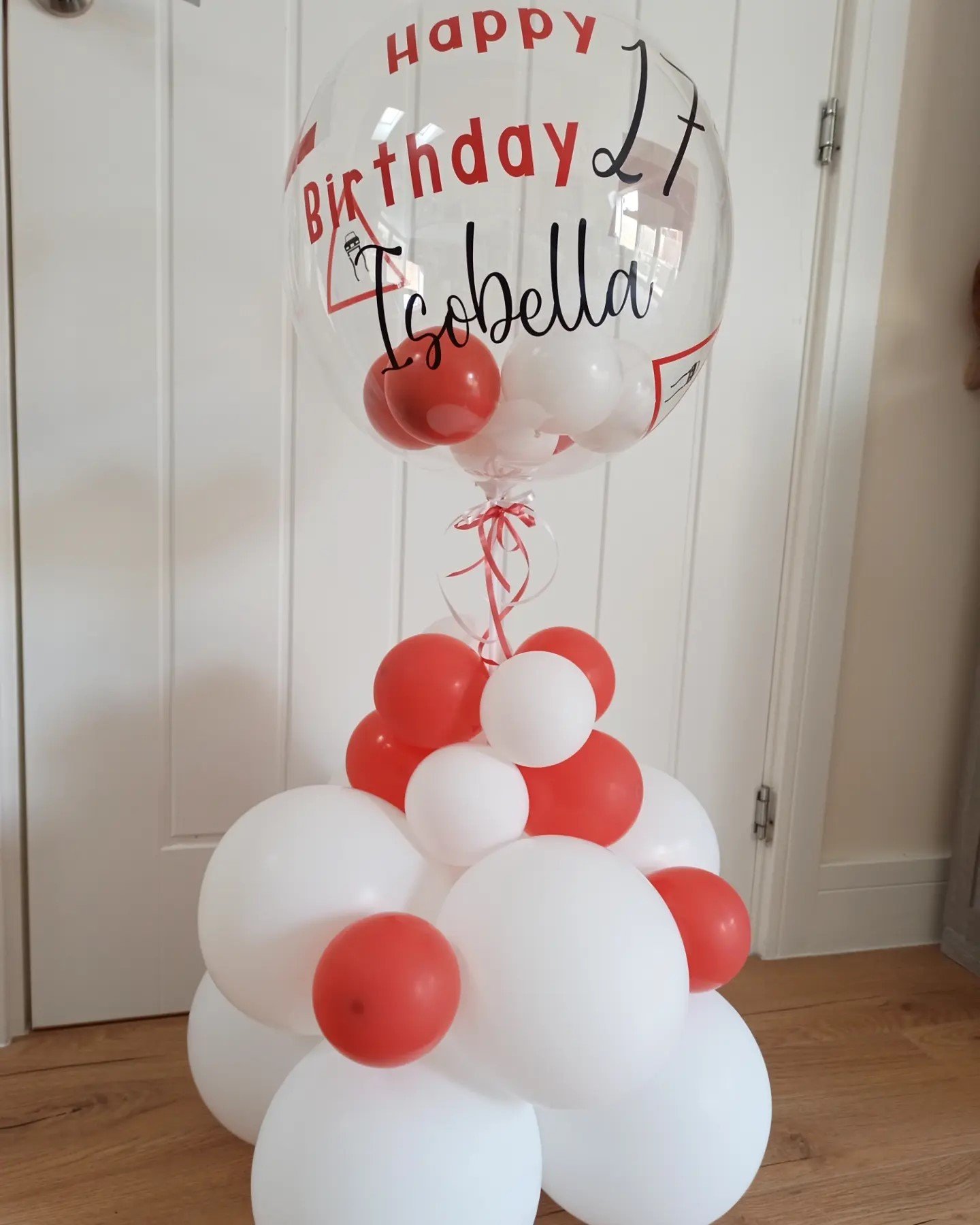 A balloon display with a large transparent balloon with red and black text that reads 'Happy Birthday Isobella', attached to a cluster of smaller white and red balloons.