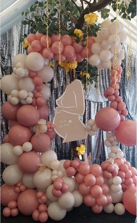 Decorative balloon arrangement with pink, white, and cream balloons, greenery, yellow ribbon accents, and a central sign in the shape of a lingerie piece that reads 'Baby shower'.