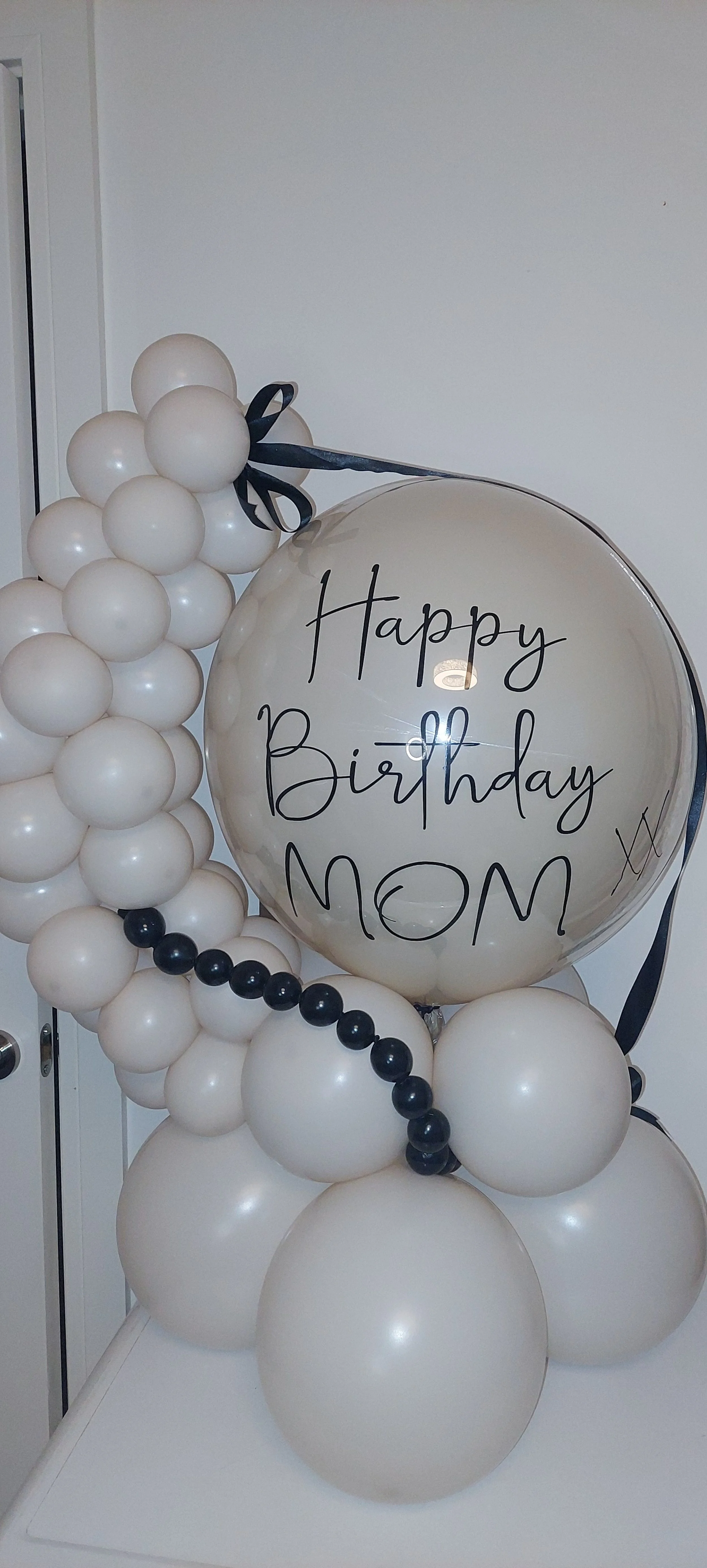 Balloon bouquet with a large clear balloon that says "Happy Birthday MOM" and smaller white and black balloons.