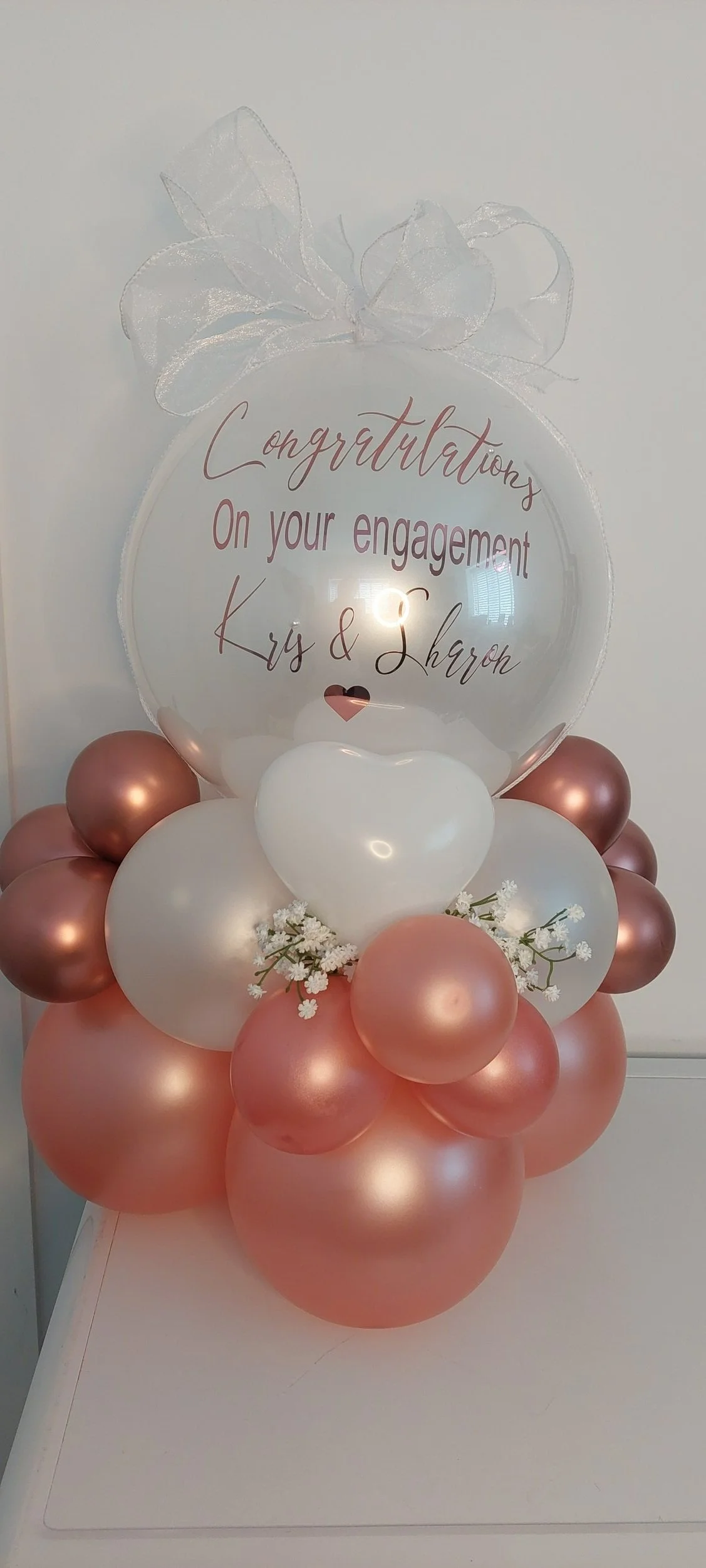 Balloon arrangement with a transparent balloon reading 'Congratulations on your engagement Kris & Sharon,' surrounded by pink, white, and metallic balloons, with small white flowers.