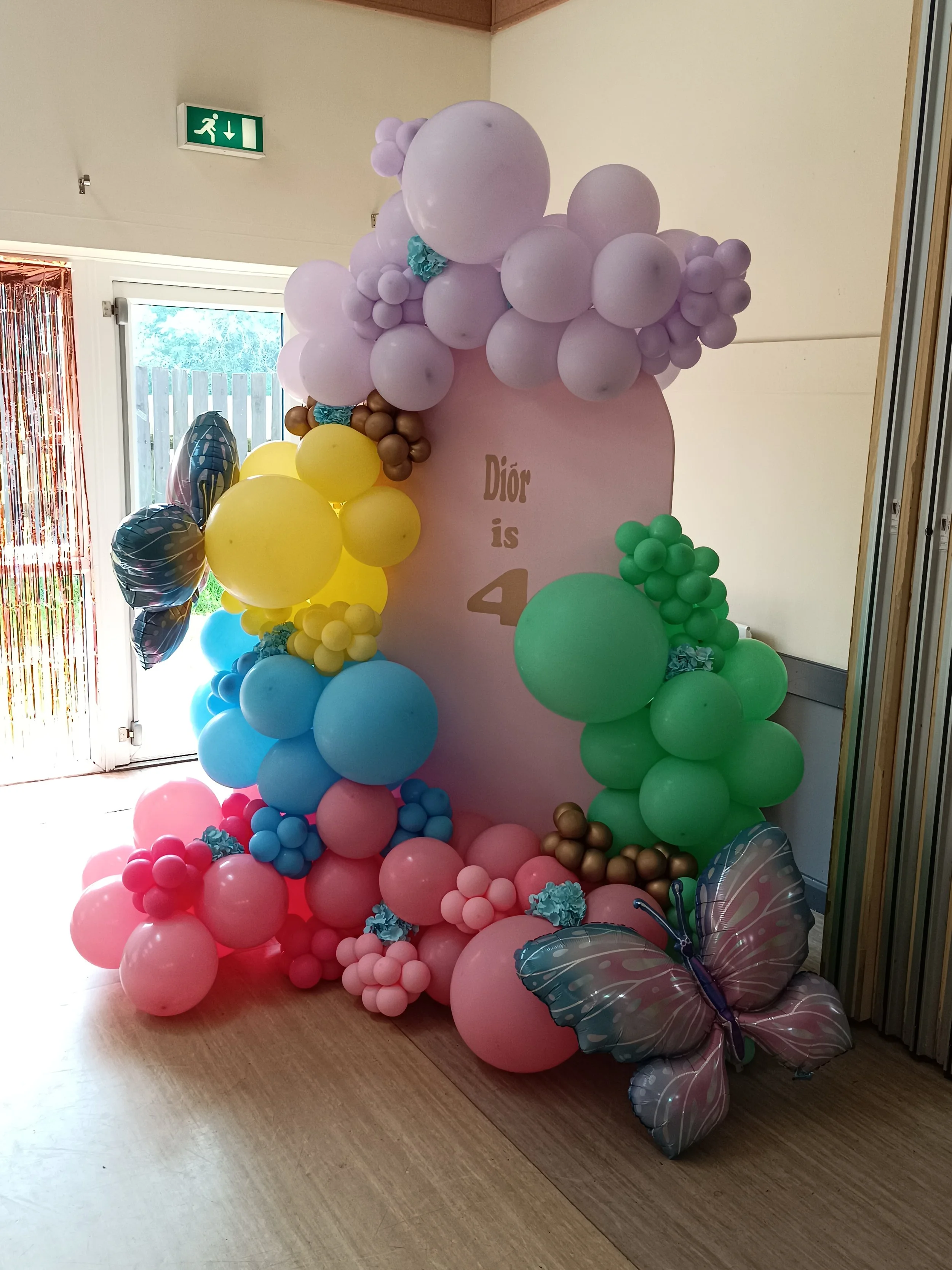 Colorful balloon arrangement with pink, purple, yellow, green, and blue balloons, some shaped like clusters and others as large single balloons, including butterfly-shaped balloons at the bottom, with a pink backdrop and partially visible decorations, for a birthday celebration.
