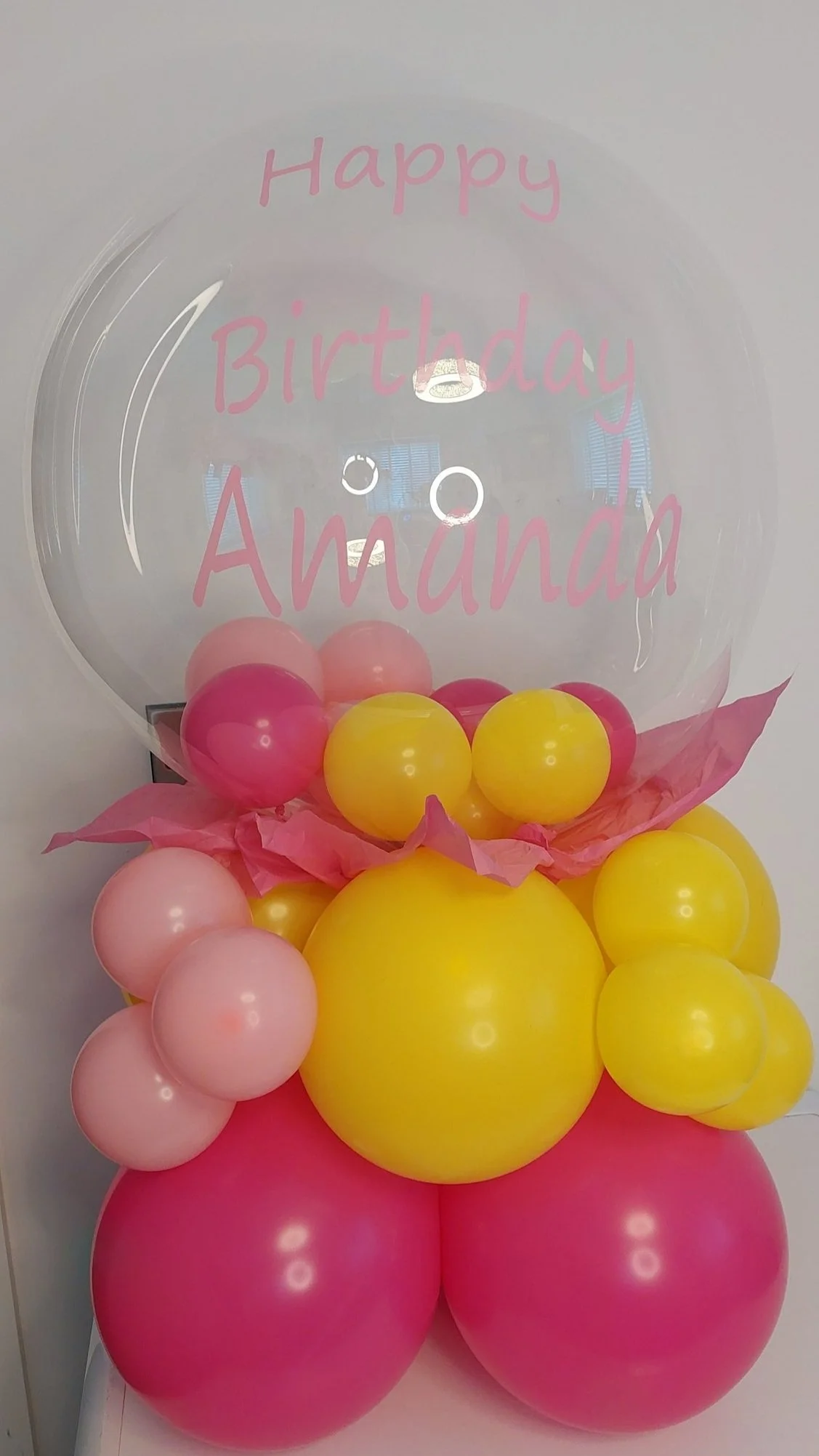 Balloon arrangement with pink, yellow, and peach balloons, topped with a clear balloon that has pink text reading "Happy Birthday Amanda."