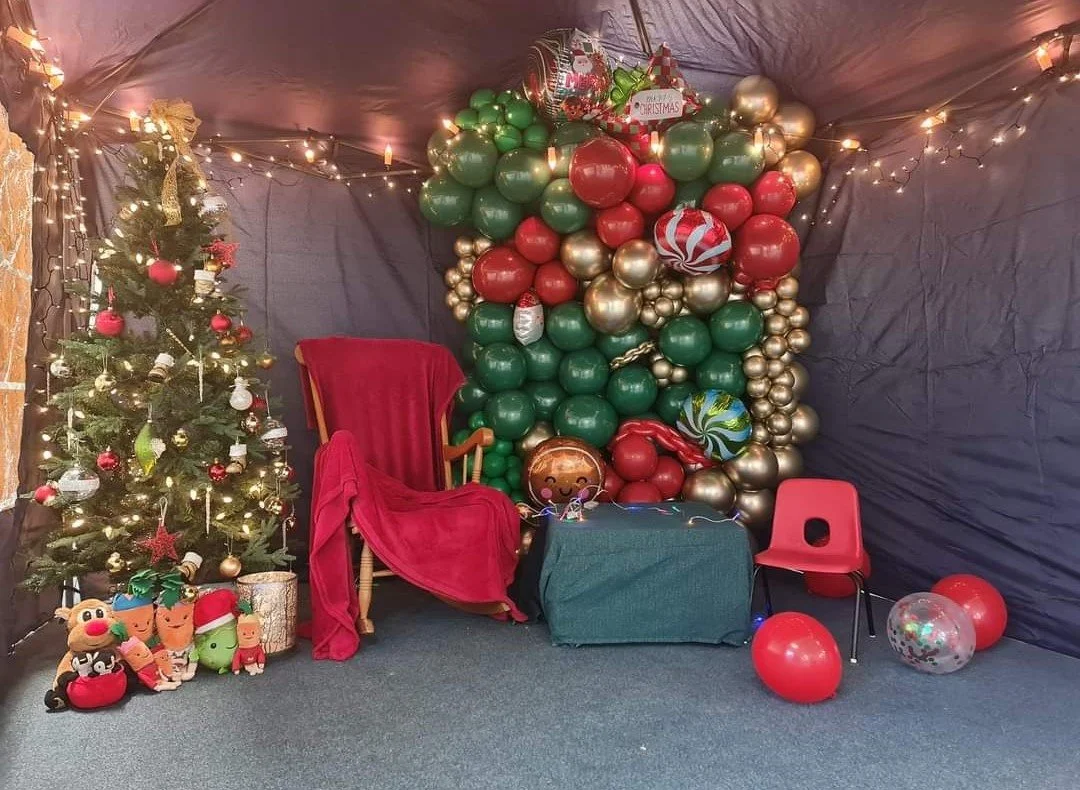 A Christmas-themed decorated room with a Christmas tree on the left, decorated with red, white, and gold ornaments and lights. There are various plush toys in front of the tree, including a reindeer, a Christmas stocking, and a green character. To the right, there is a large balloon display with red, green, gold, and patterned balloons, along with a balloon that says 'Merry Christmas'. There is a red chair, a red velvet covered wooden chair, a small table with a green cloth, and some balloons on the floor. The room has black fabric walls and is decorated with string lights.