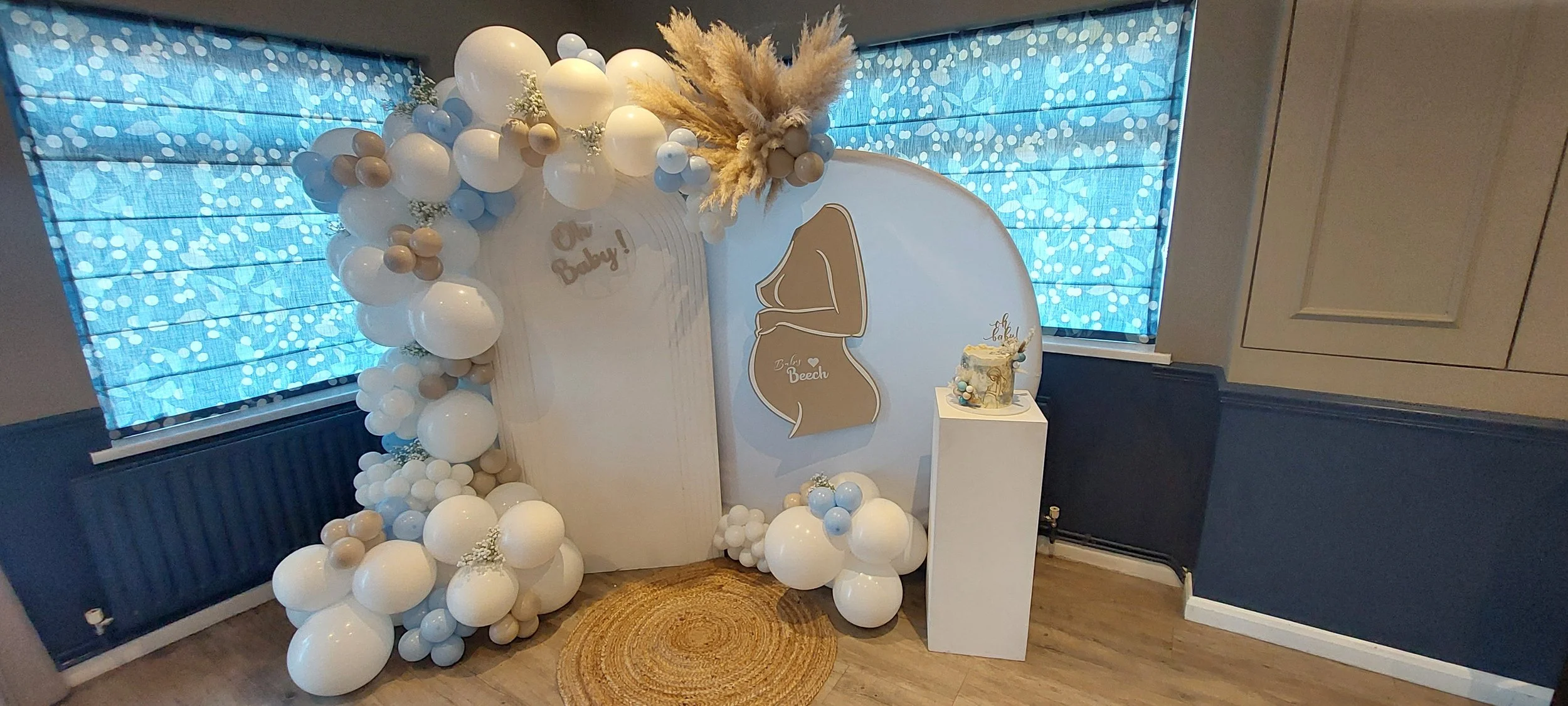 Beach-themed baby shower decoration with white and beige balloons, a small cake on a white pedestal, a large white backdrop with a beach outline and the words "Baby Beech," and windows with blue patterned blinds.
