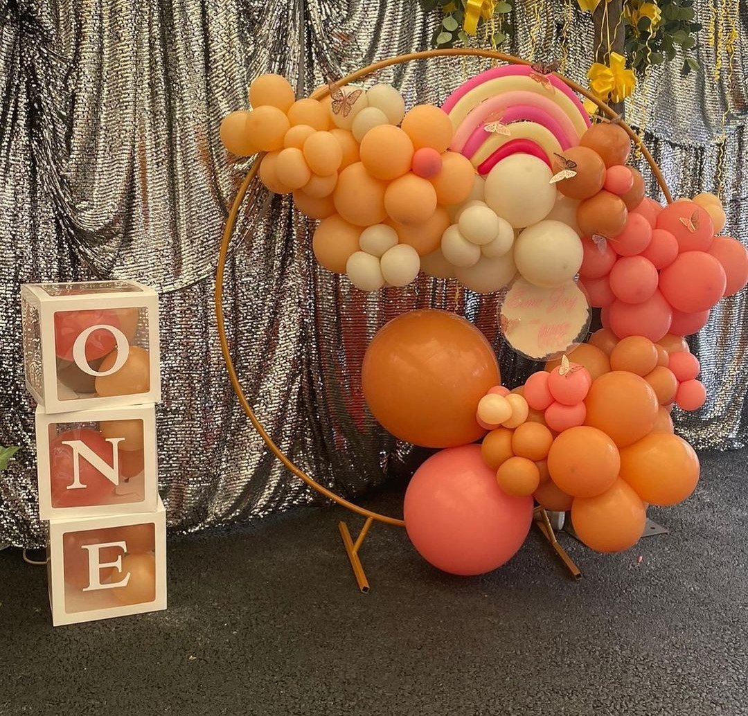 Decorative balloon arrangement with pastel pink, peach, white, and gold balloons, arranged in a circle-like display with a rainbow and butterfly accents. To the left, a white block with the letters O, N, and E spelled out with peach-colored balloons inside the boxes. Silver sequin backdrop.
