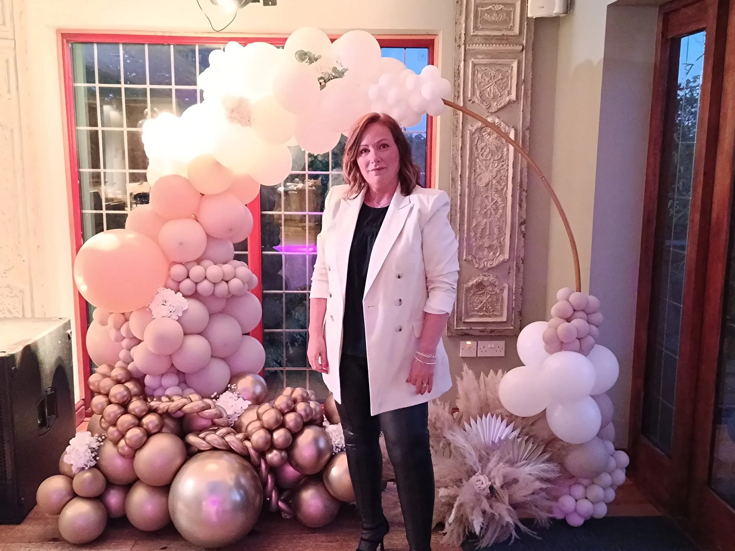 Woman standing in front of a decorative balloon arch with soft pink, gold, and white balloons, and a window with red frame in the background.