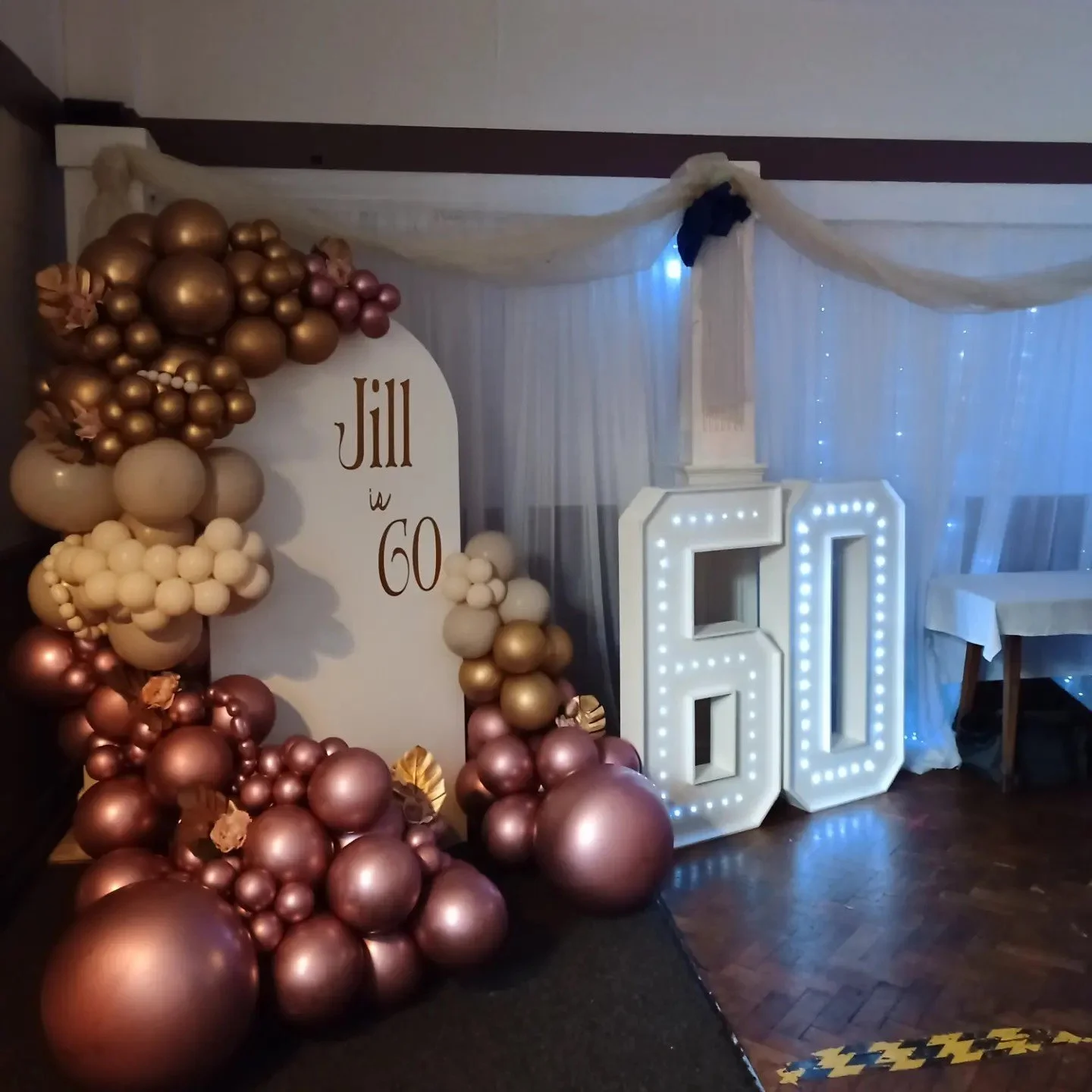 Decorative setup for Jill's 60th birthday celebration featuring a balloon arrangement in shades of pink, cream, and gold, a white sign with 'Jill is 60' written on it, illuminated giant white number '60', and sheer white curtains with blue lighting in the background.