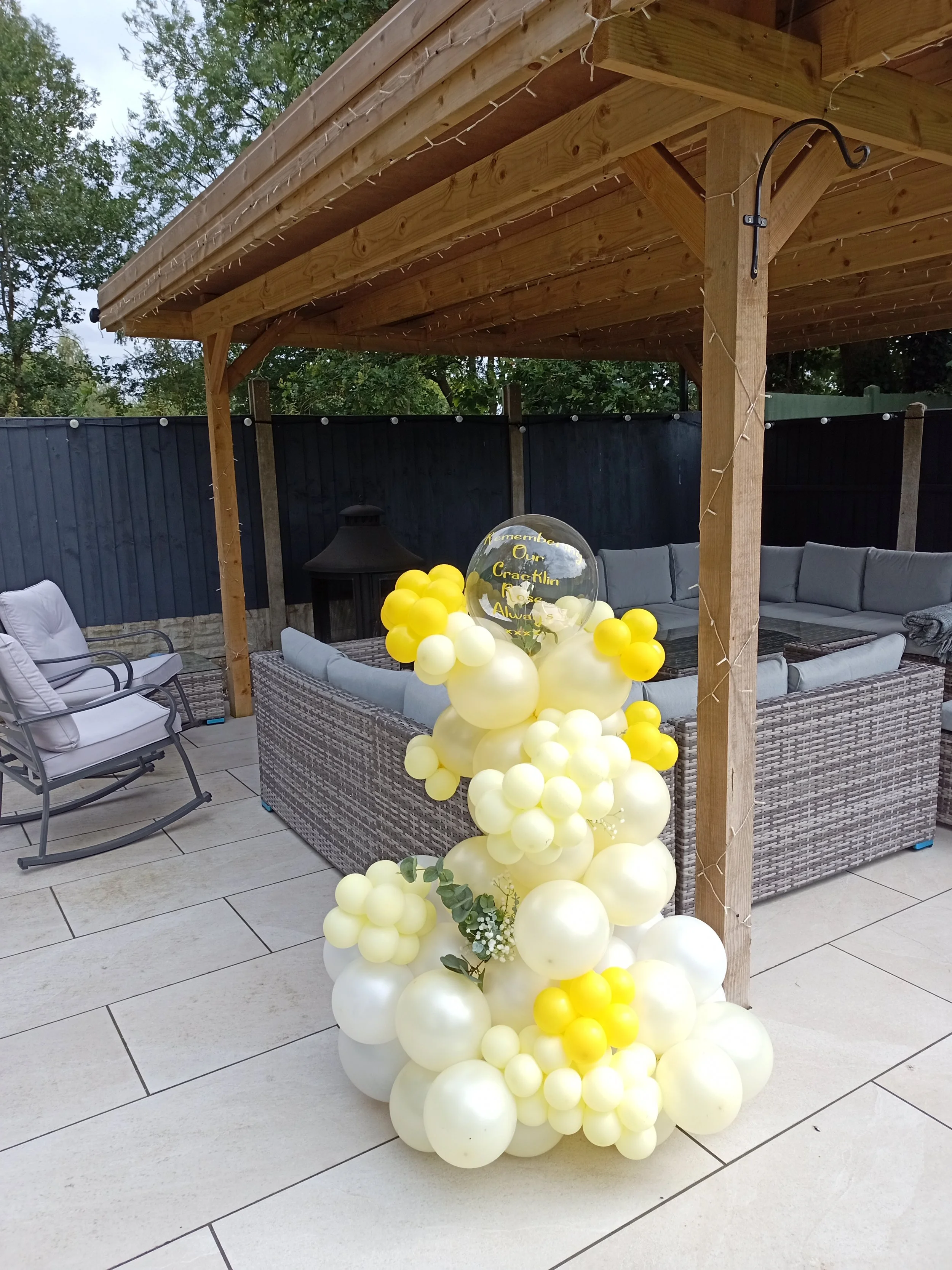 Decorative yellow and white balloons with a clear acrylic balloon in the center that reads 'Remember Our Cracklin Rose Always' on a patio with outdoor furniture and a wooden canopy