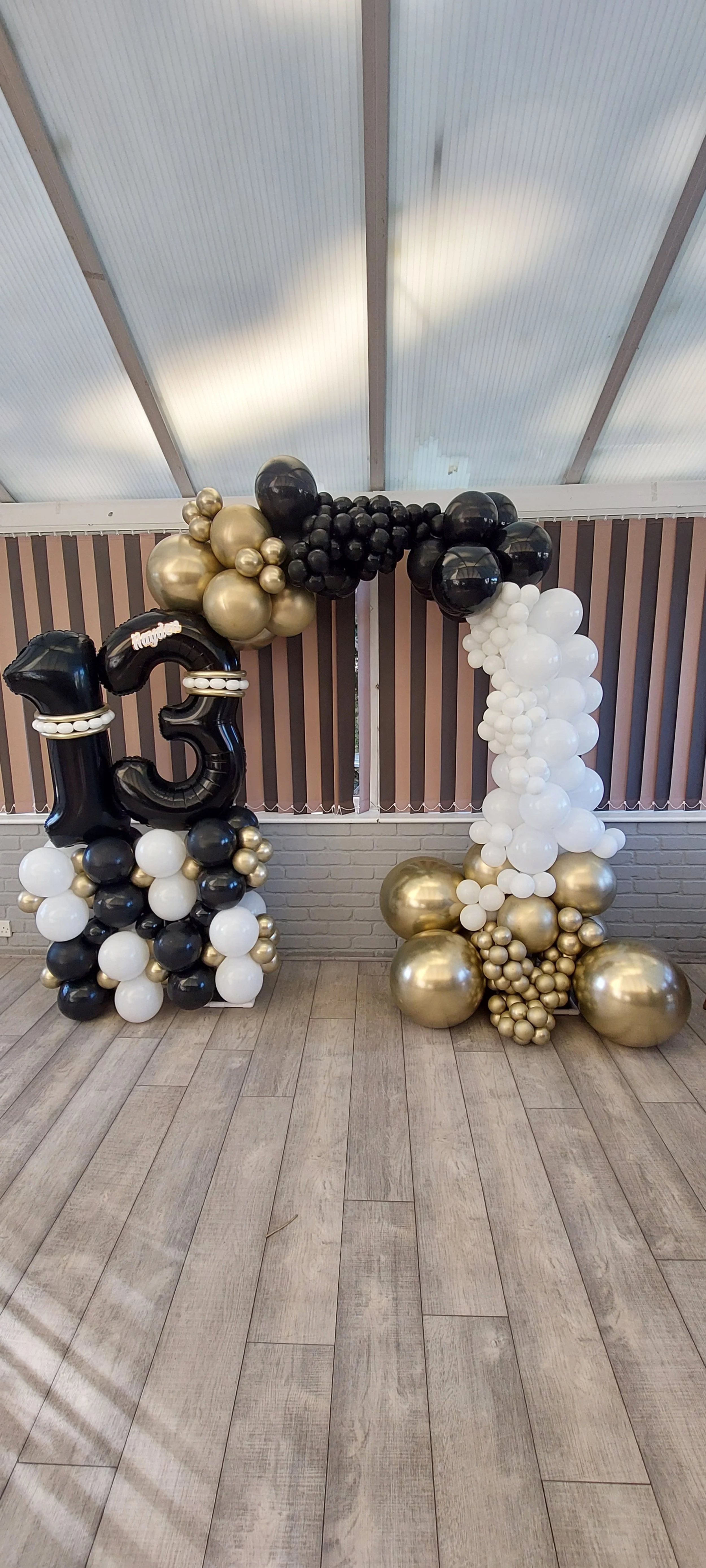 Balloon arch with black, white, gold, and pearl balloons, including a large black '15' balloon, set against a backdrop with vertical blinds and wooden flooring.