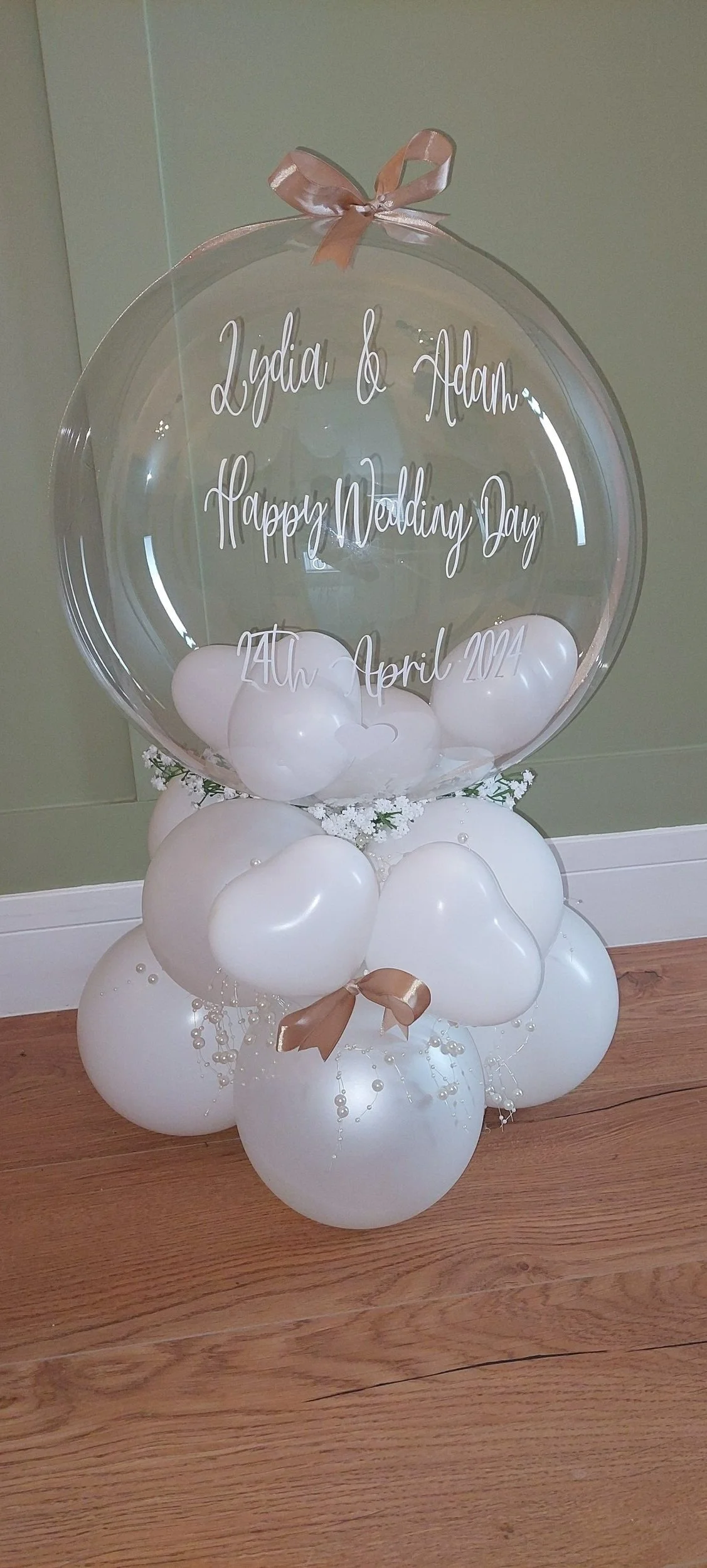 Decorative balloon arrangement with a large clear balloon on top, inscribed with white cursive text reading "Lydia & Adam Happy Wedding Day 24th April 2024". The arrangement includes white heart-shaped balloons, small white flowers, bows, and pearl-like beads.