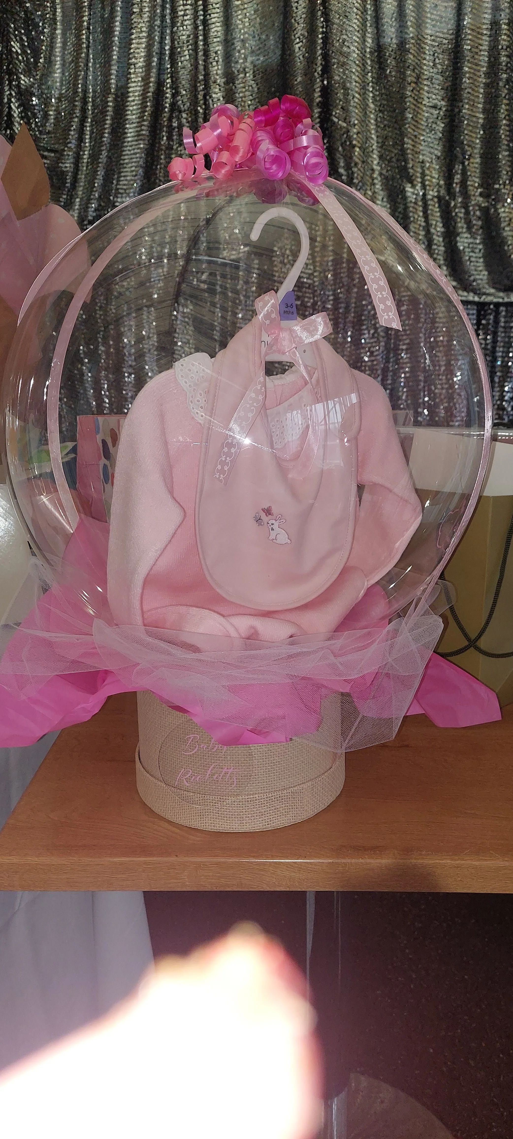 Pink baby outfit with a bib, displayed inside a transparent case with pink decorations, on a wooden surface.