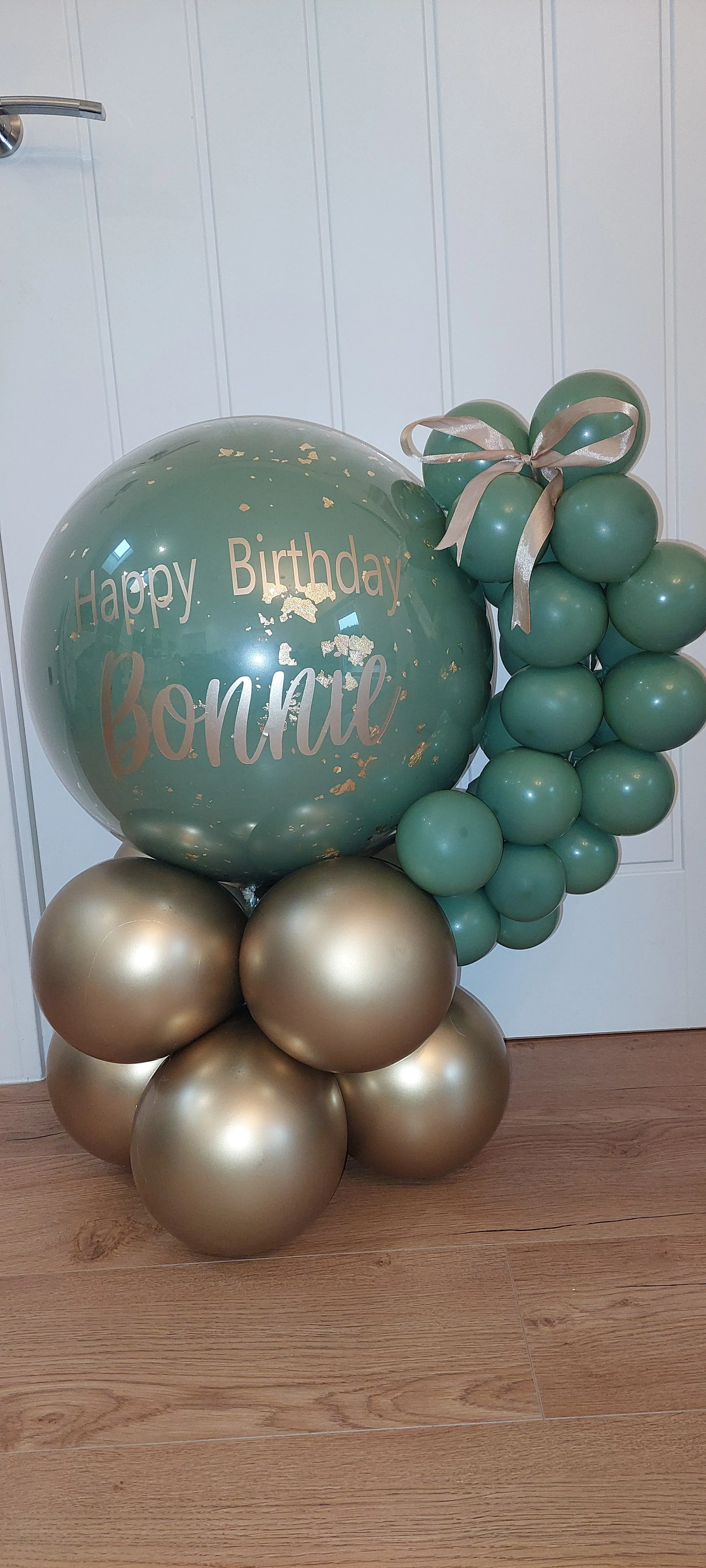 Green and gold balloon arrangement with a large green balloon that says 'Happy Birthday Bonini' and a cluster of smaller green balloons, tied with a gold ribbon, set against a white paneled wall.