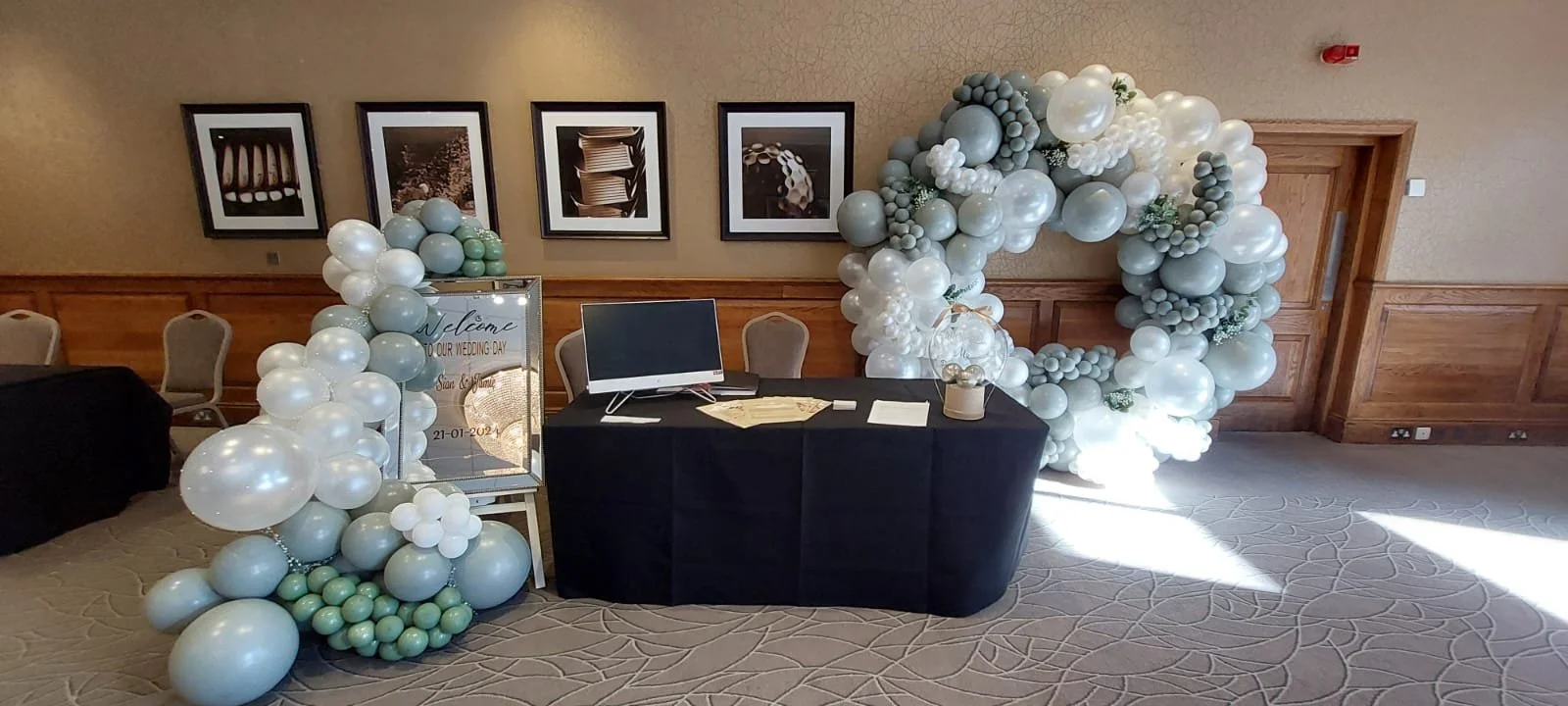 Balloon arch and decorations at a wedding venue, with a welcome sign, table, and framed pictures on the wall.