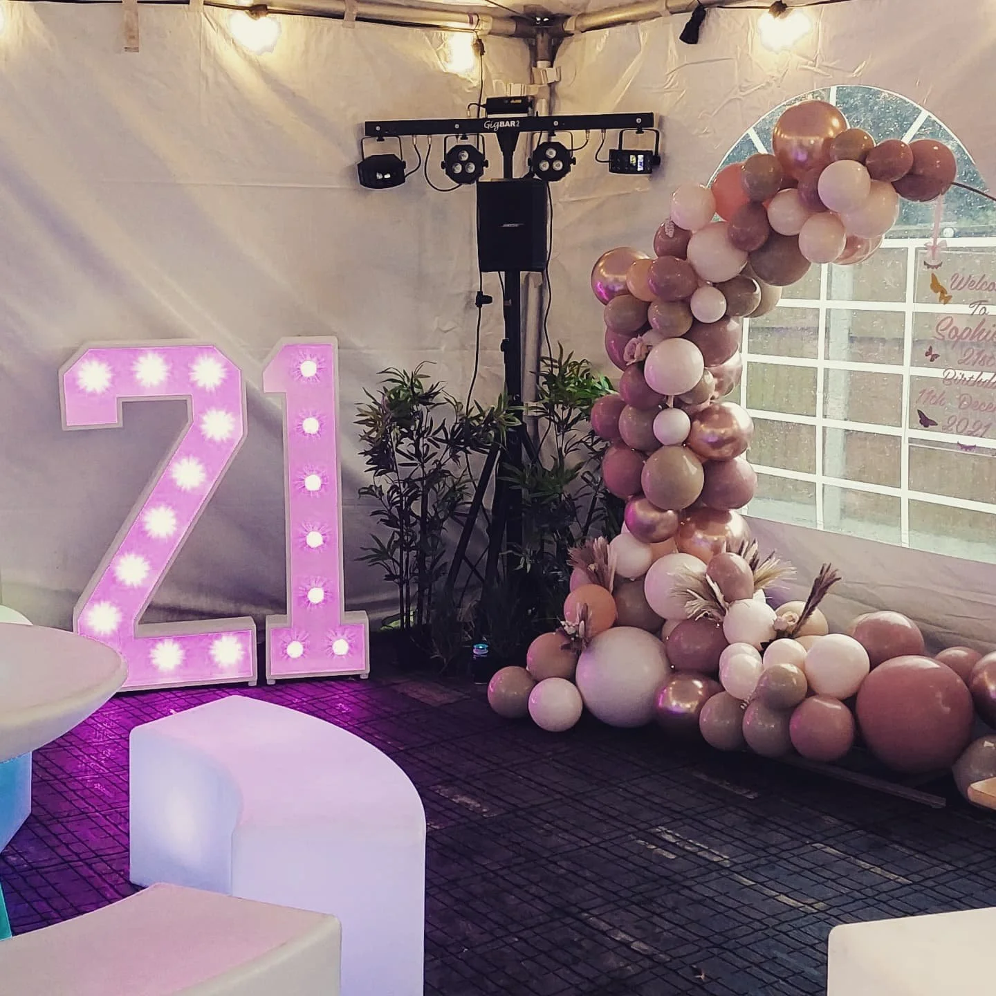 Decorative event setup with illuminated number 21, pink and neutral-colored balloon arrangement, and DJ equipment in an enclosed space.
