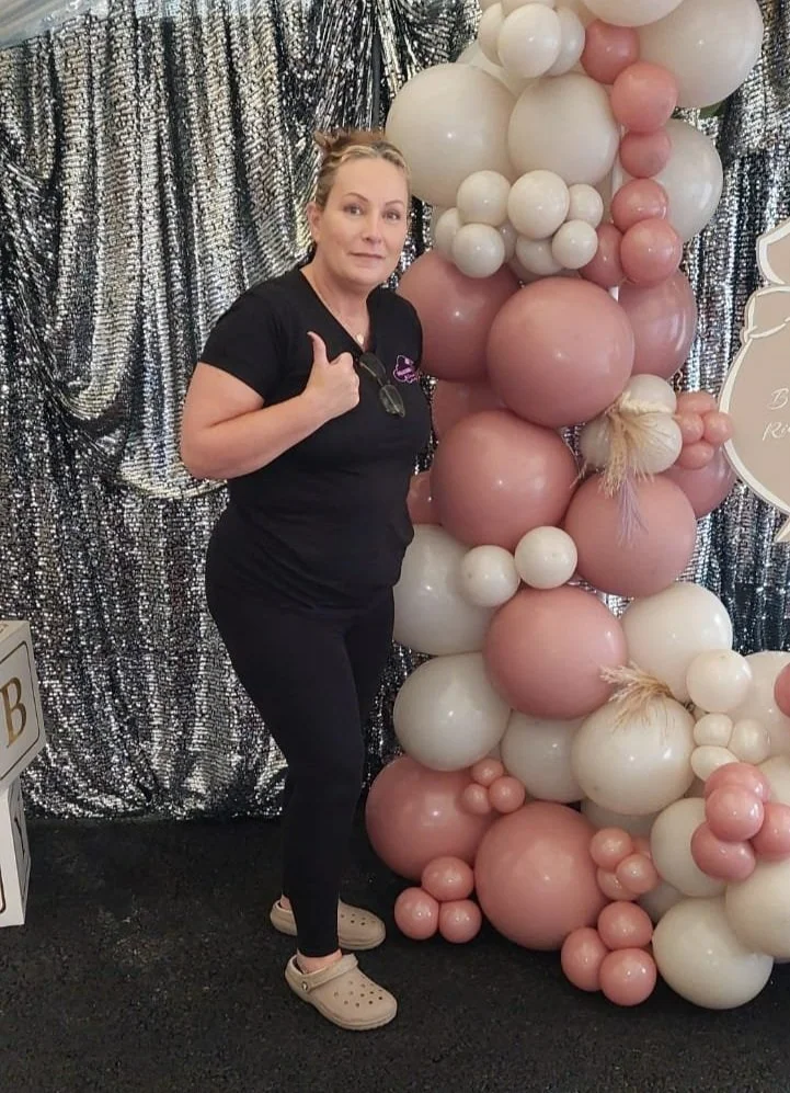 Woman in black shirt and pants giving a thumbs-up next to a large balloon sculpture made of pink, white, and peach-colored balloons, with a metallic sequin backdrop.