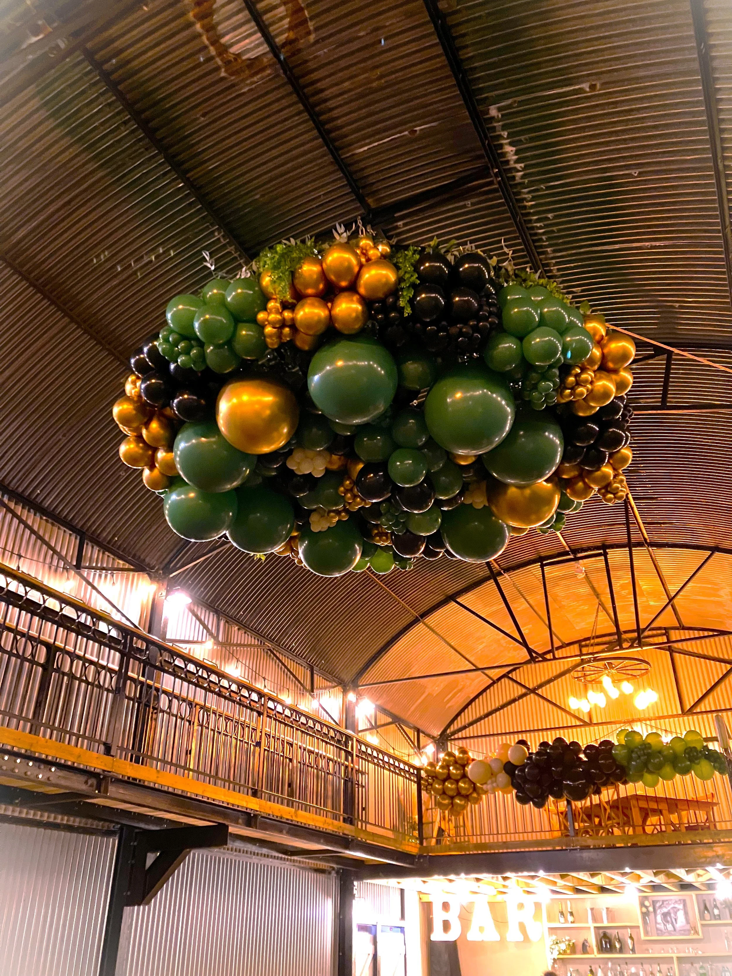 Decorative balloon arrangement with black, gold, green, and white balloons hanging from a metal ceiling inside a venue or bar.