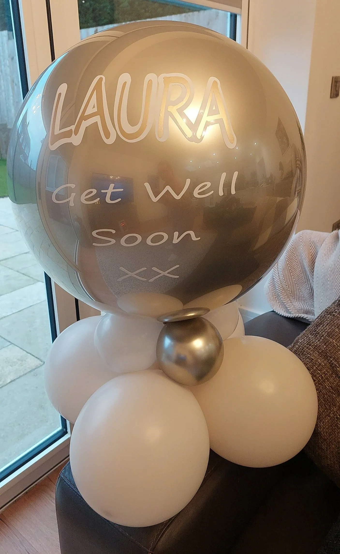 A cluster of white and gold balloons, with a silver balloon in the center, attached to a large reflective balloon that reads 'LAURA Get well soon xxx', placed near a window with a view of a backyard.