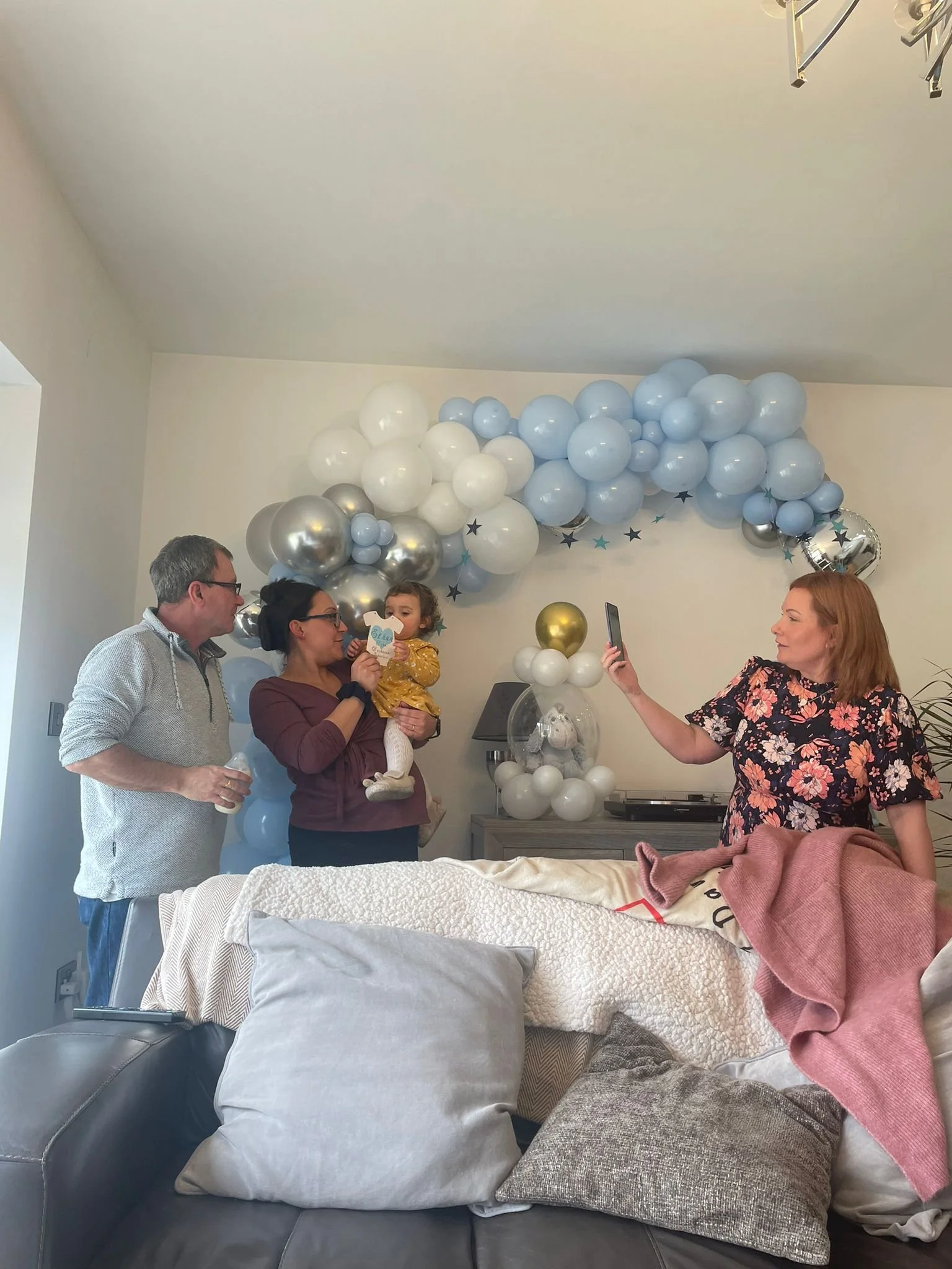 People gathered in a room decorated with a balloon arch and stars, with one person taking a selfie, as others hold a young girl, likely celebrating a special occasion.