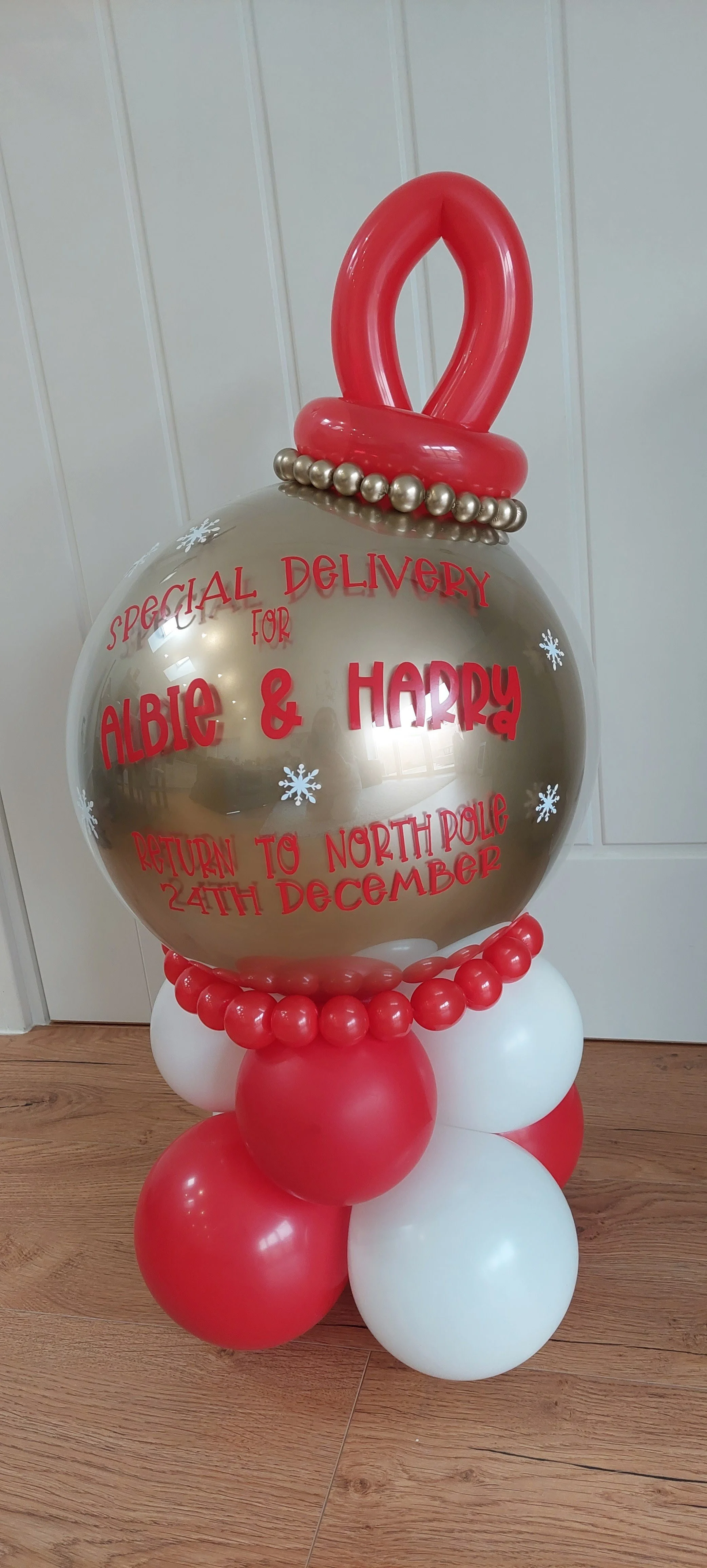A balloon decoration resembling a Christmas ornament with a gold sphere on top, red and white balloons at the base, and red lettering for a Christmas delivery message.