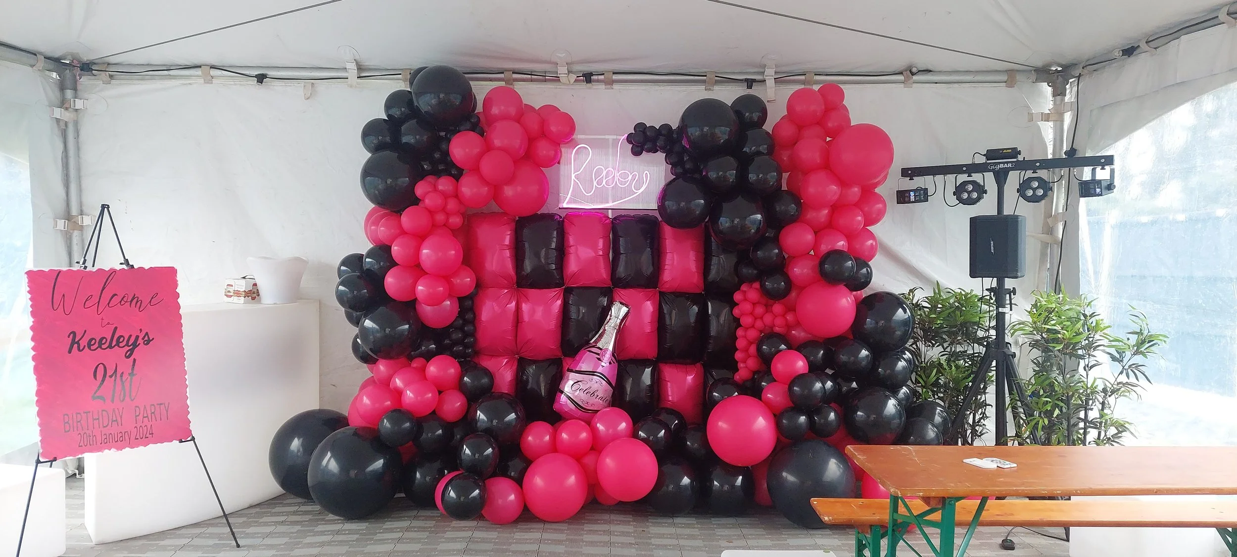 Balloon display with pink and black balloons and a neon sign that reads 'Party'. There is a pink sign that says 'Welcome Keeley's 21st Birthday Party, 20th January 2024' on a stand to the left, and DJ equipment with speakers to the right.