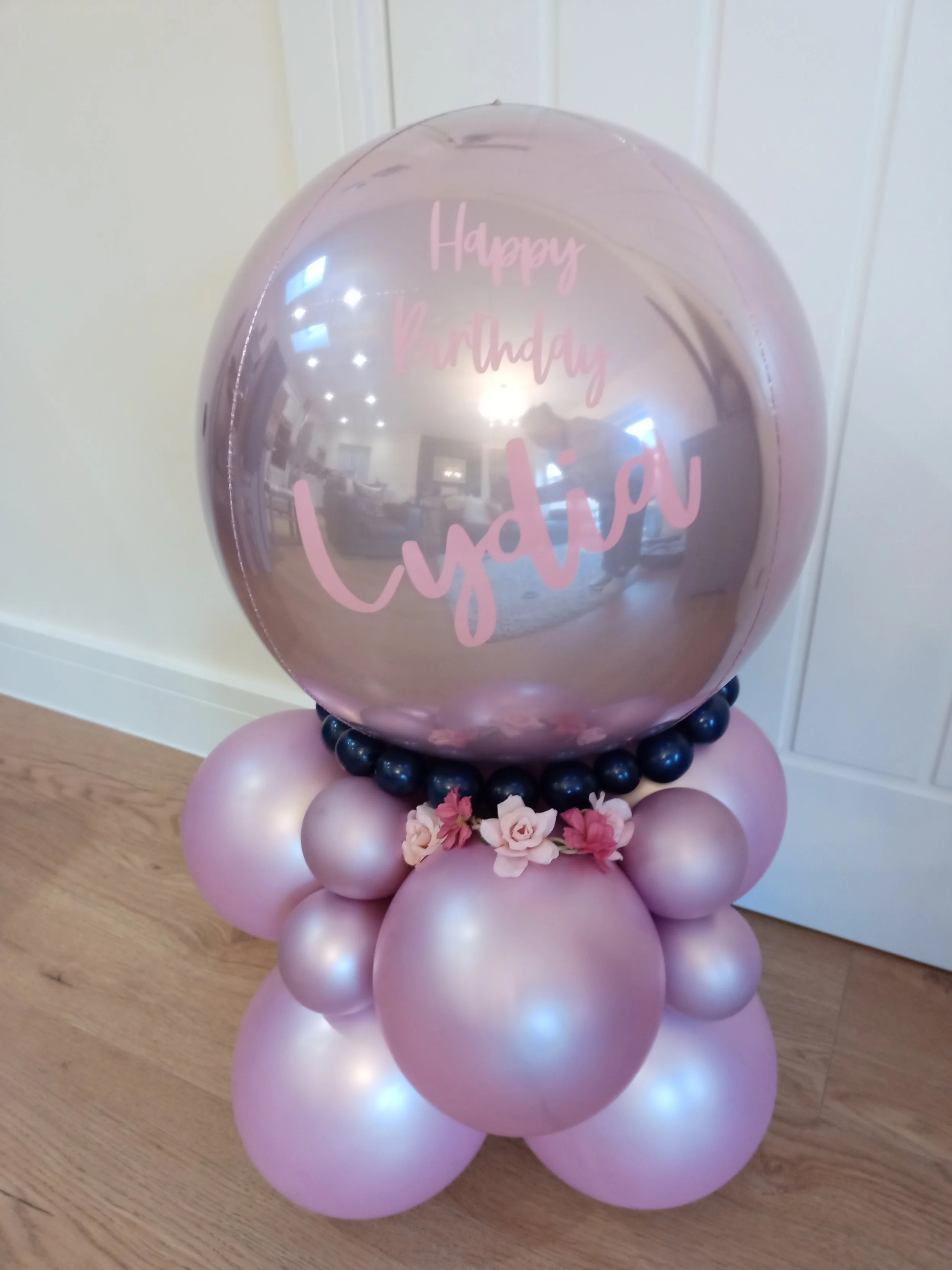 A balloon display with a large pink metallic balloon that has 'Happy Birthday' written on it, surrounded by smaller pink and black balloons, and decorated with pink and white flowers at the base.