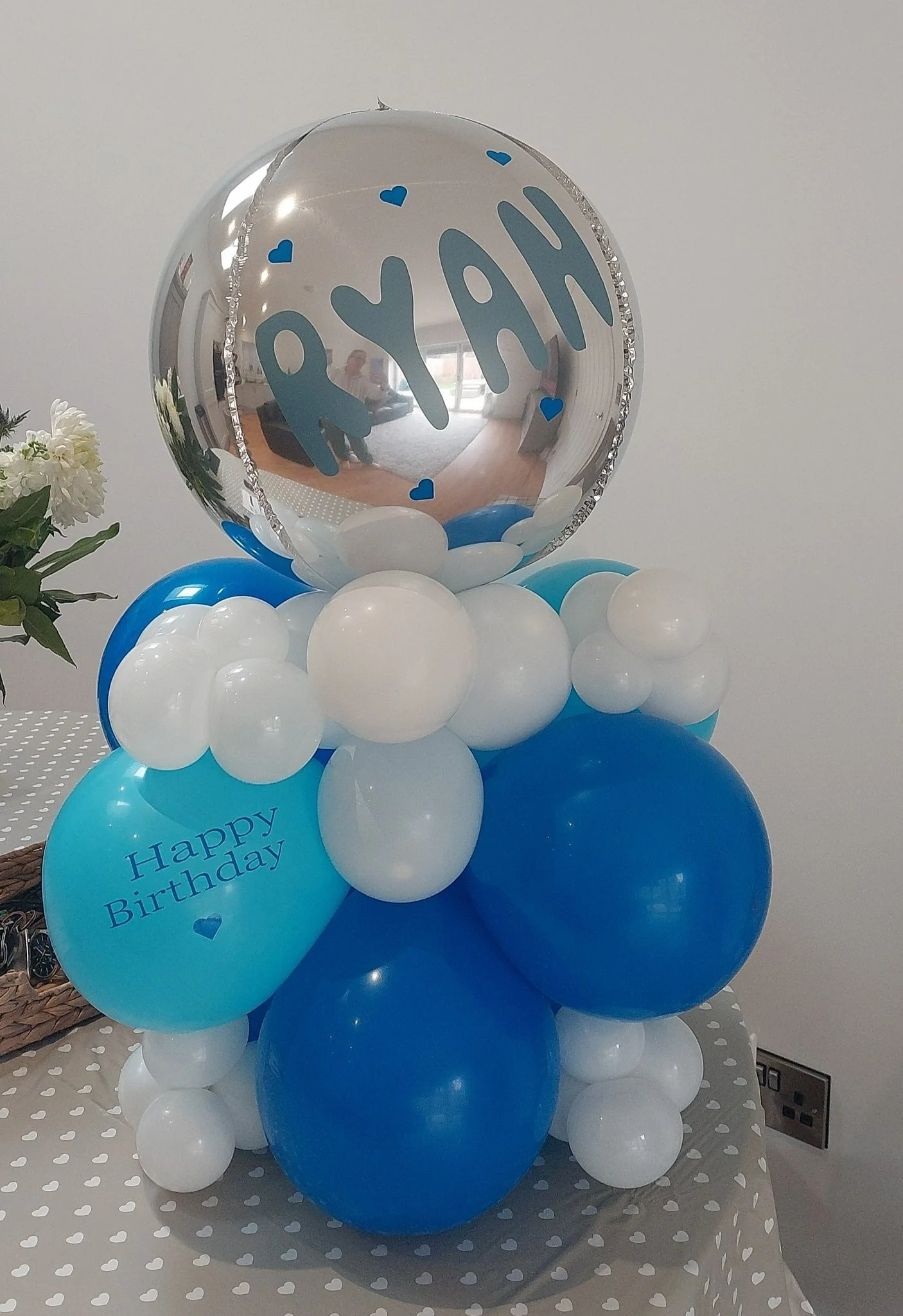 A large silver balloon with the word "YAH" written on it in blue, surrounded by smaller blue and white balloons, with a blue balloon that has "Happy Birthday" written on it in blue, all arranged in a decorative display.