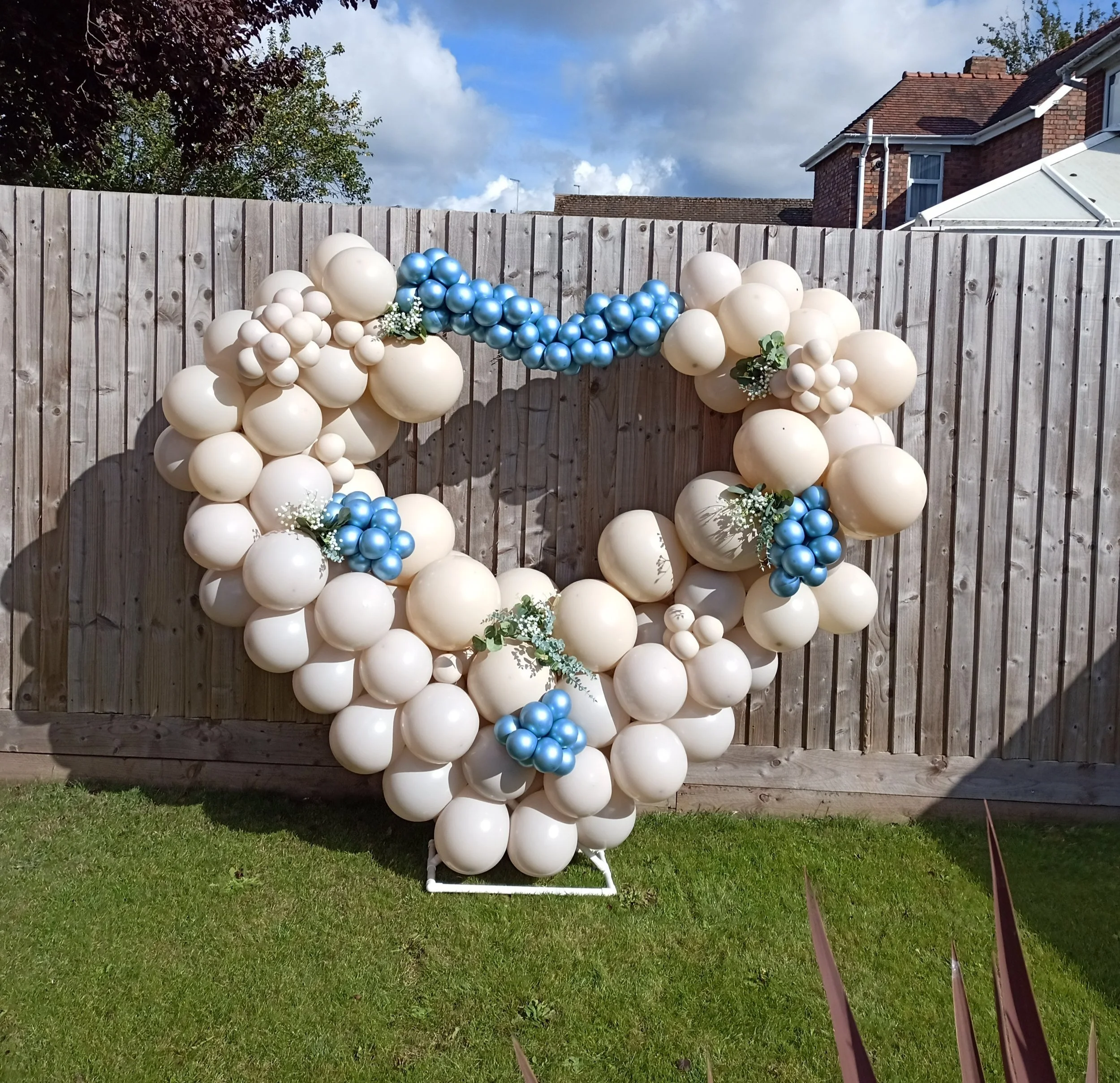 Decorative balloon arch with white and blue balloons, adorned with small white flowers, set up outdoors against a wooden fence with houses and a cloudy sky in the background.