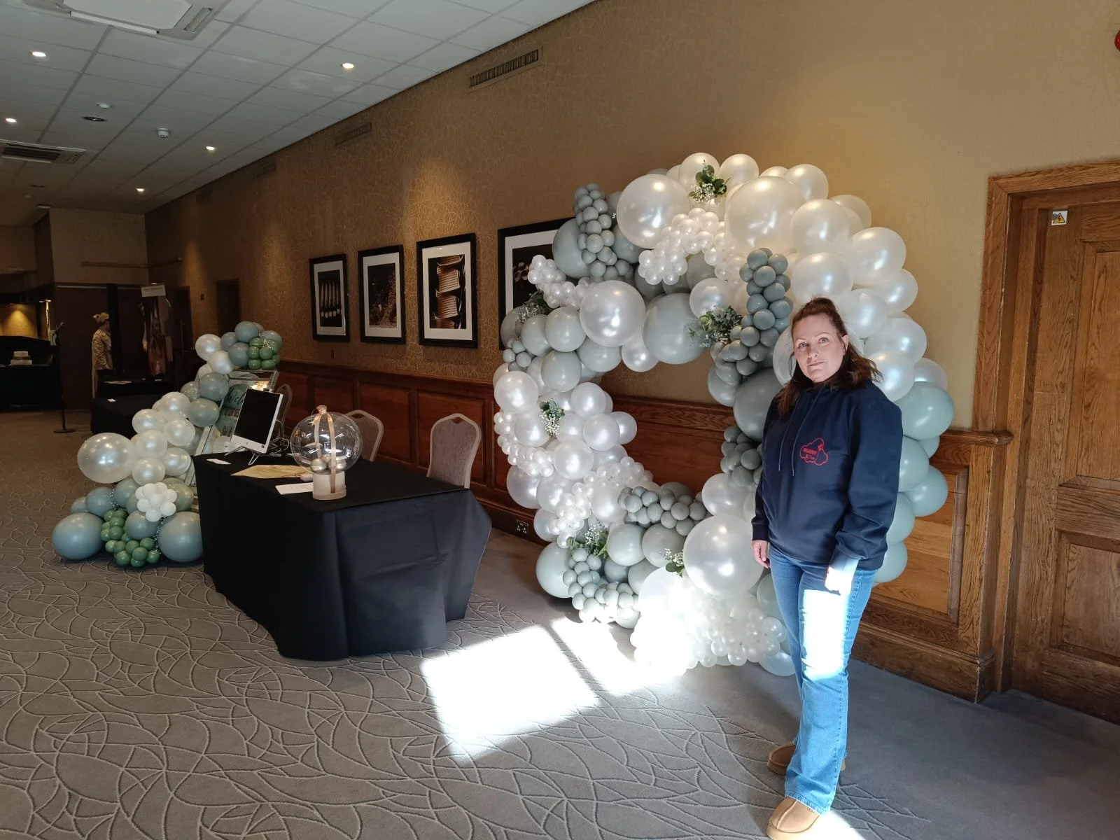 A woman standing in front of a white balloon arch decorated with gray and white balloons at an indoor event space.