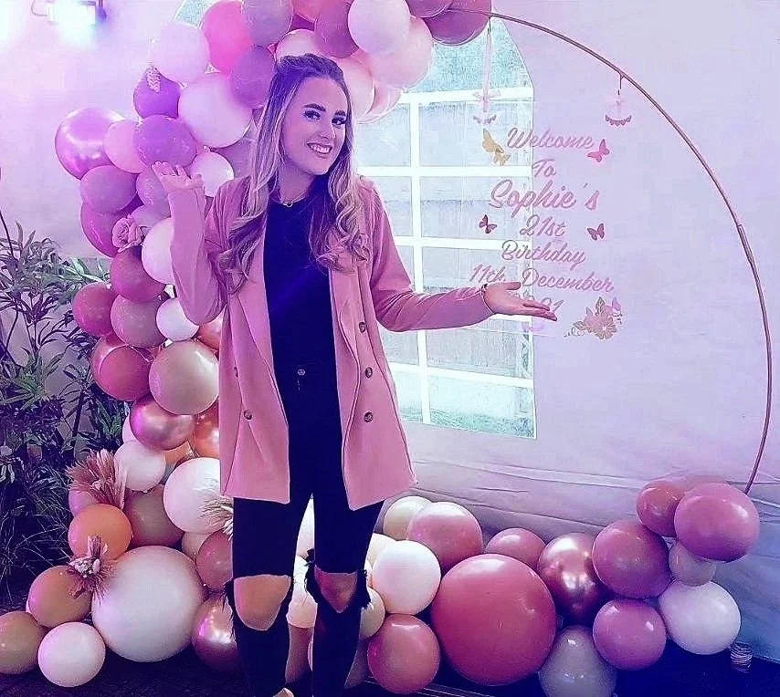 Young woman standing in front of a pink balloon arch and a birthday sign, celebrating Sophie’s 16th birthday on December 11, 2023.
