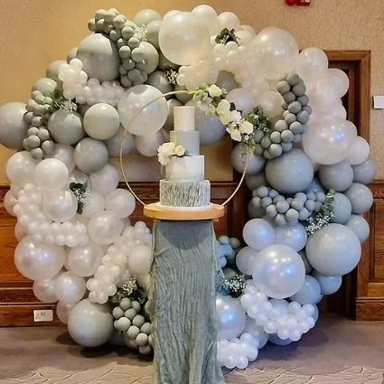 A decorative balloon wreath with white and gray balloons surrounding a wedding cake on a wooden stand, in front of a wooden wall.