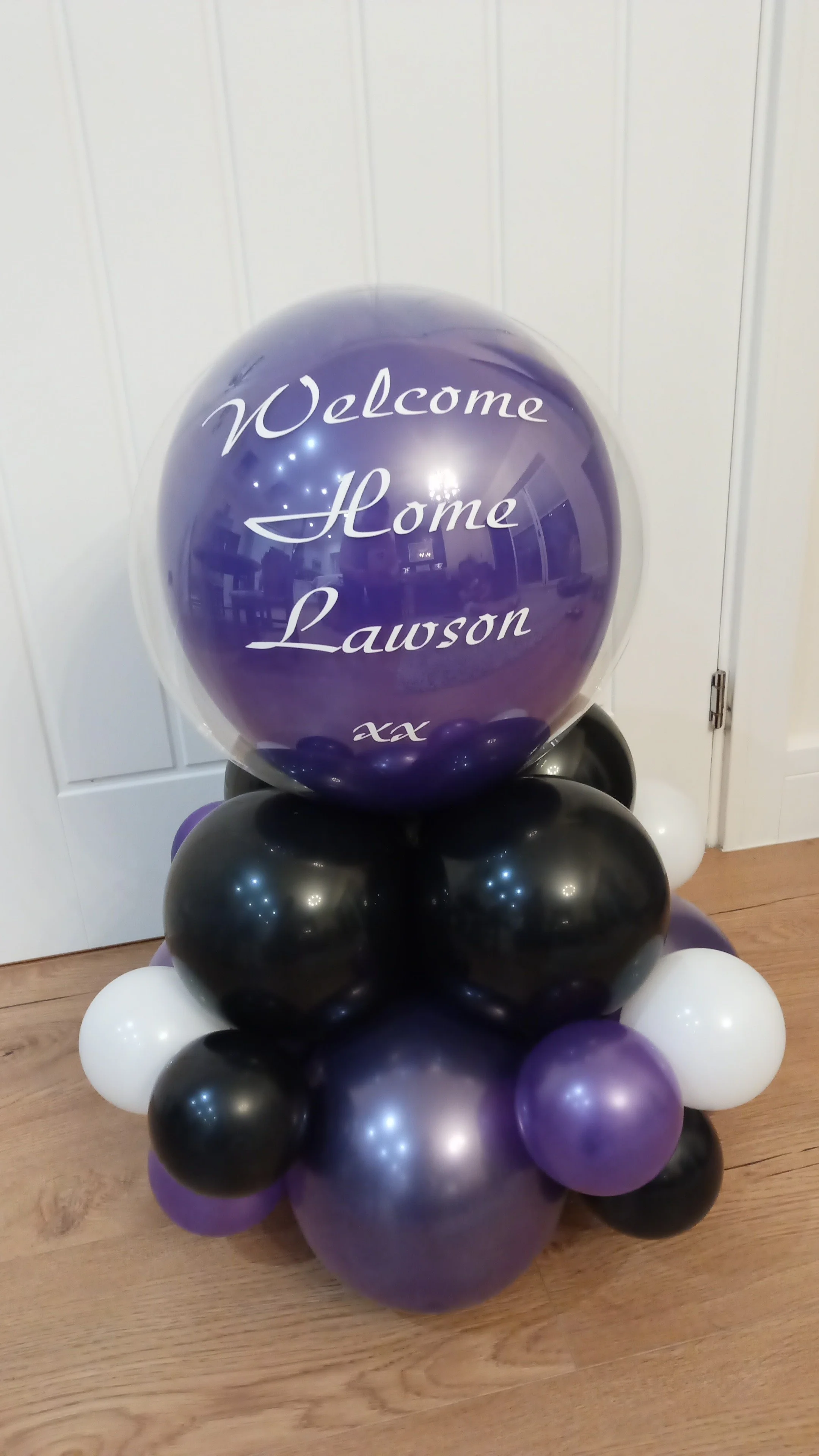 Balloon arrangement with a large purple balloon that has 'Welcome Home Lawson xx' written on it, surrounded by smaller black, white, and purple balloons.