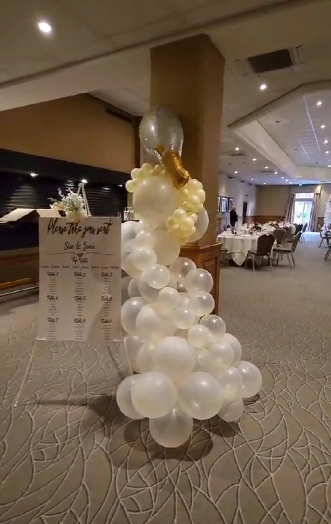 Balloon decoration with an arrangement of white balloons and a large champagne bottle balloon on top, next to a sign-in or guestbook table in a banquet hall.