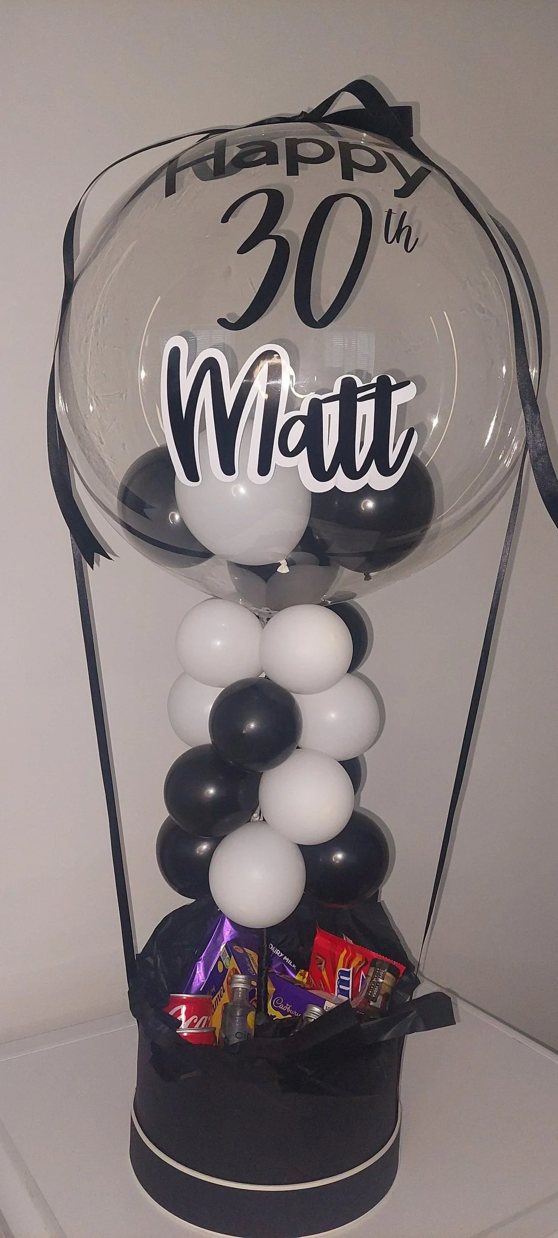 A decorative balloon bouquet for a 30th birthday celebration, featuring a large transparent balloon with black and white balloons inside, and a black and white balloon cluster at the base with a gift bag containing chocolates.