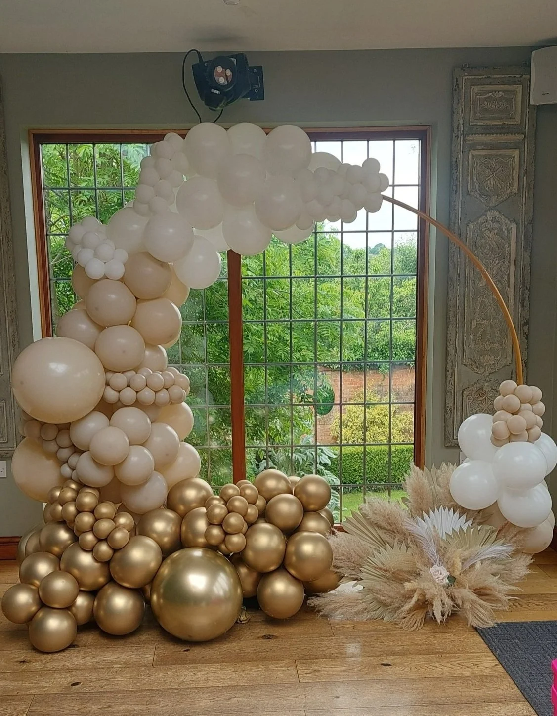 Balloon arrangement with white, beige, and gold balloons next to a large window, with a decorative dried floral arrangement on the floor.
