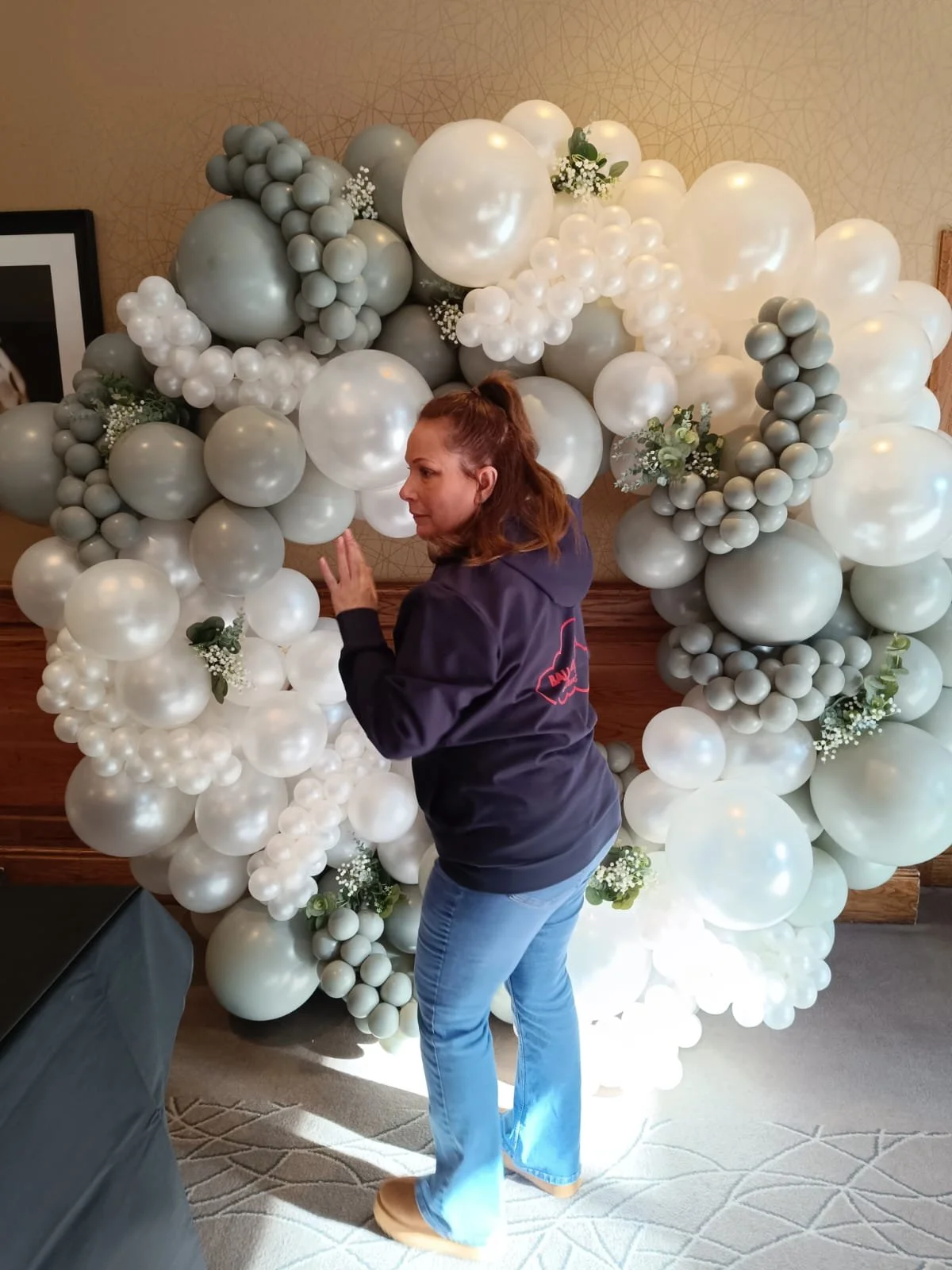 Woman standing next to a large, decorative balloon arrangement with white, gray, and pearl-colored balloons and small floral accents in front of a textured wall.