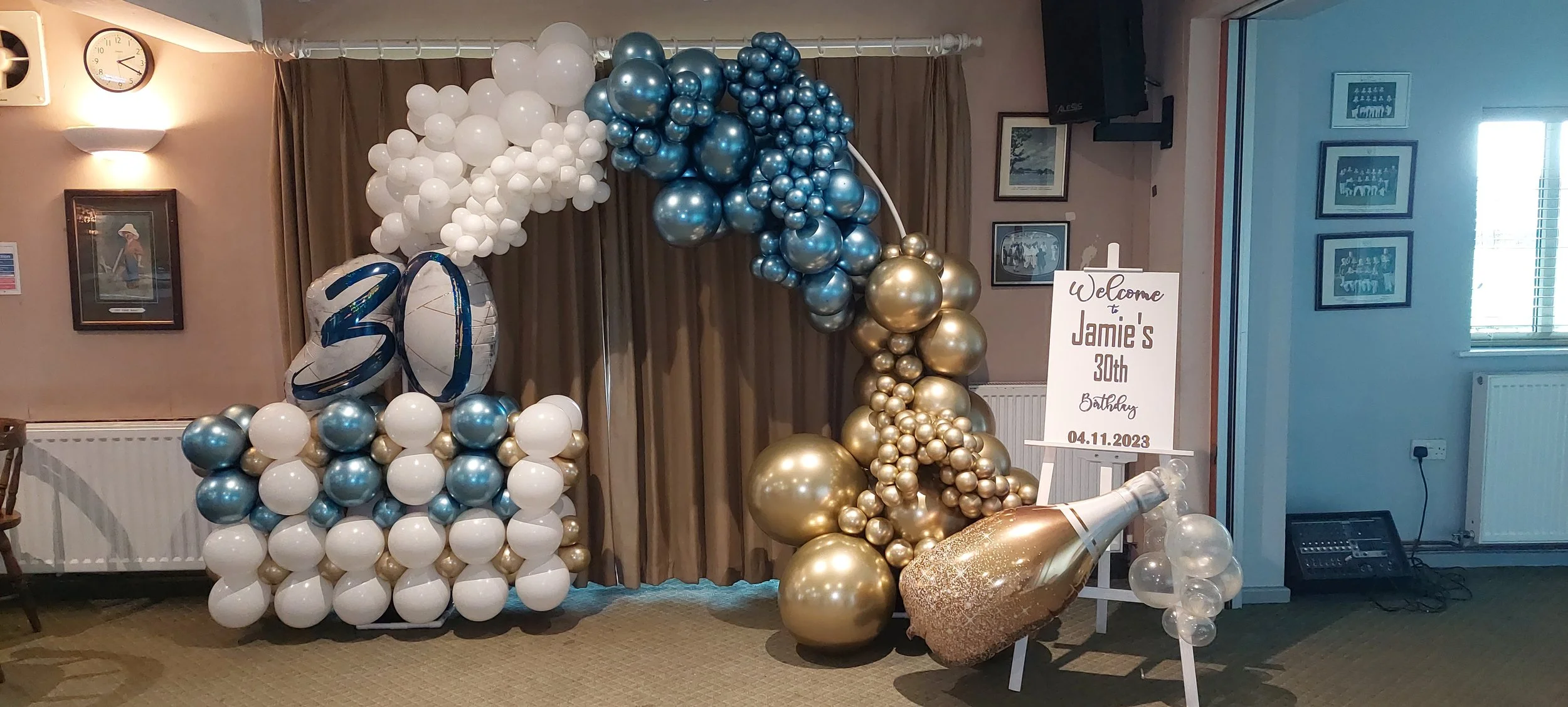Balloon arch with white, blue, gold, and silver balloons, a large gold champagne bottle decoration, and a sign that reads "Welcome to Jamie's 30th Birthday 04.11.2023" at an indoor celebration.