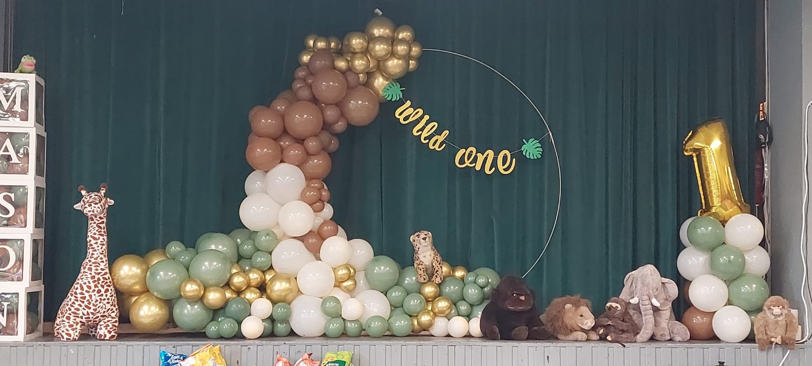 Decorations for a first birthday celebration with balloons, plush animal toys including a giraffe, cheetah, elephant, lion, and monkey, and a sign that reads 'wild one' in gold lettering.
