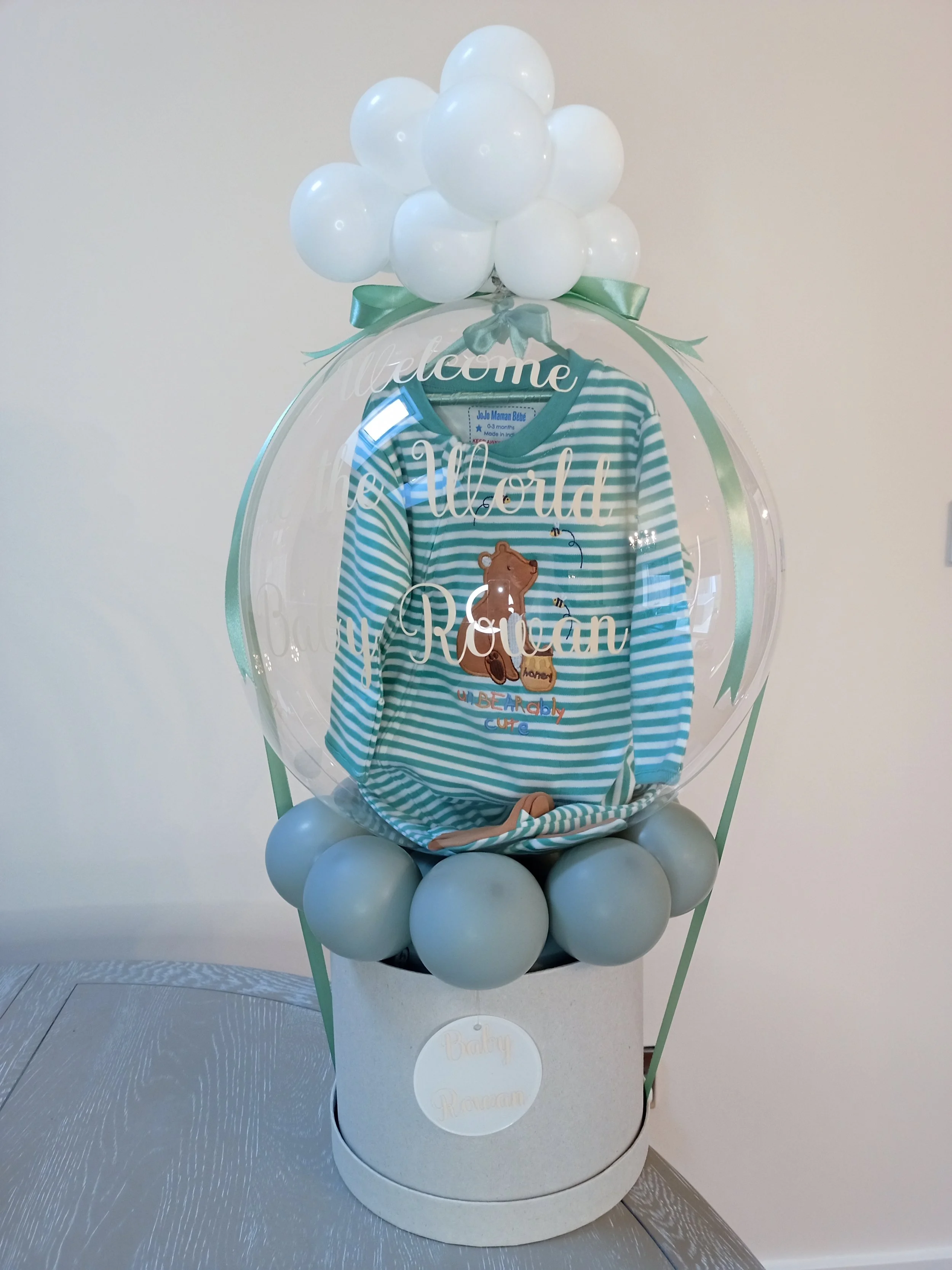 Balloon arrangement of white balloons on top of a clear spherical display with a light green ribbon, featuring a baby onesie with stripes, a bear image, and the words 'Welcome Baby Rowan' on the transparent surface.