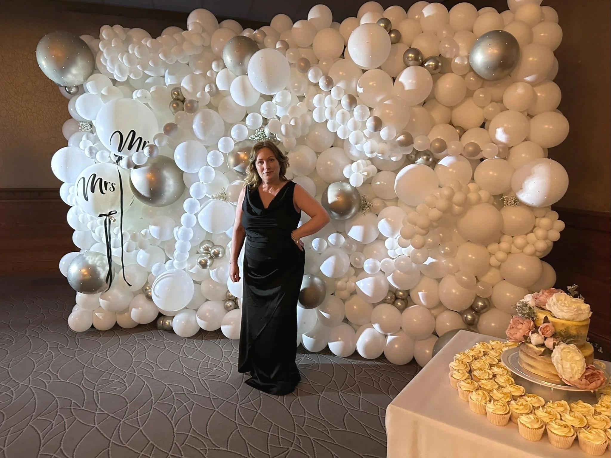 A woman in a black gown posing in front of a large white balloon display at an event. The display includes various sizes of white and silver balloons with two balloons featuring the words 'Mr.' and 'Mrs.' written on them. There is a table on the right side with cupcakes and pink roses.