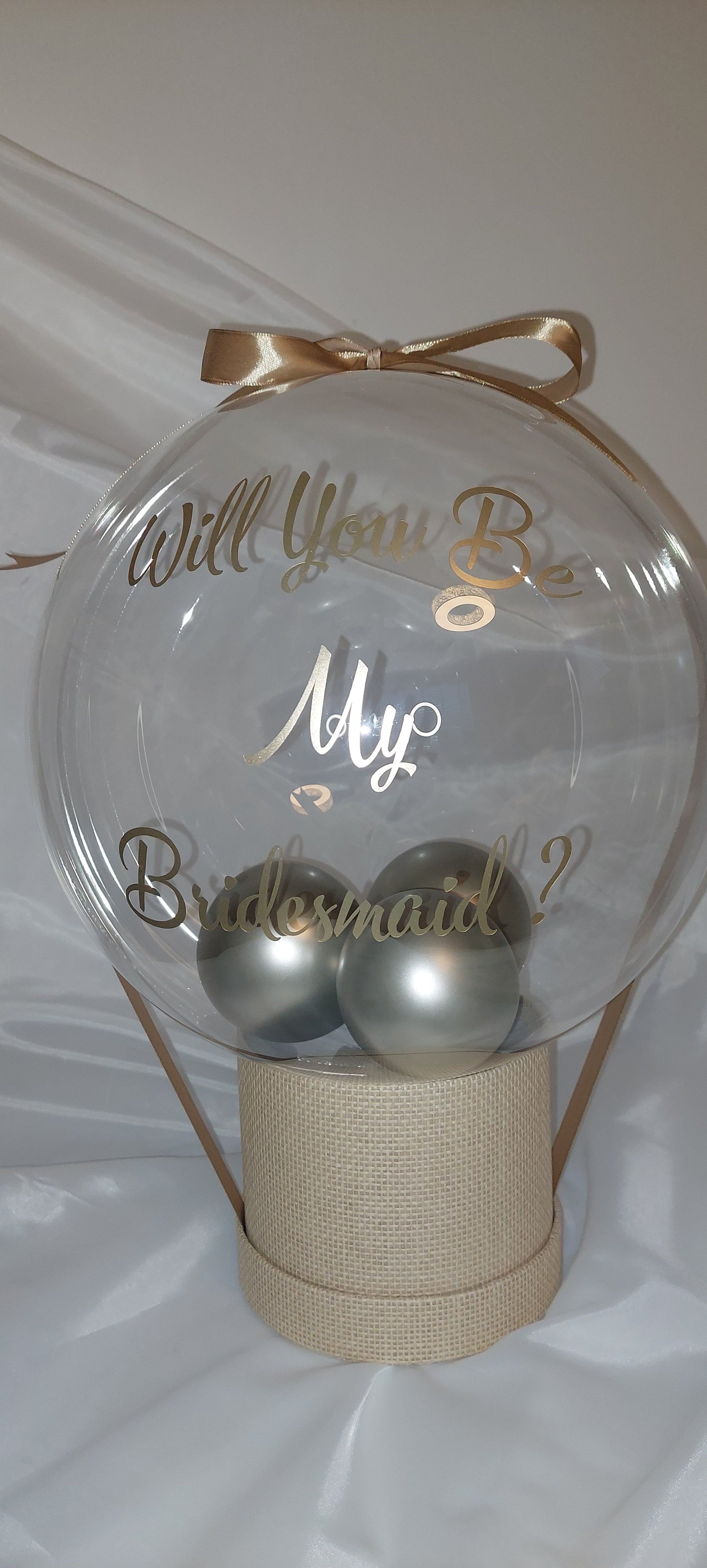 A clear balloon with gold text reading, 'Will You Be My Bridesmaid?', attached to a beige ribbon and base, with three silver decorative balls inside.