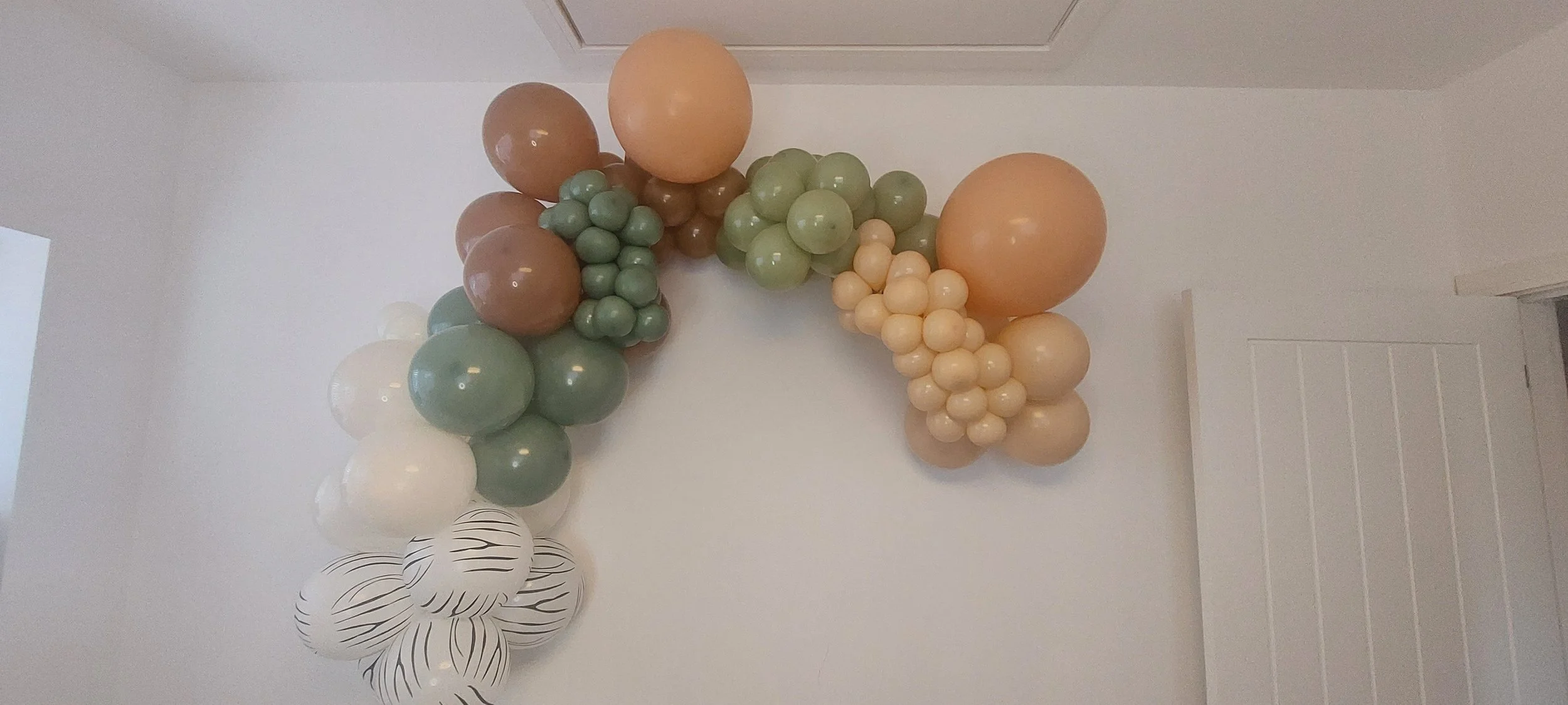 Decorative balloon arrangement with various sizes and colors, including peach, green, brown, white, and striped black-and-white balloons, attached to a white wall near a door.