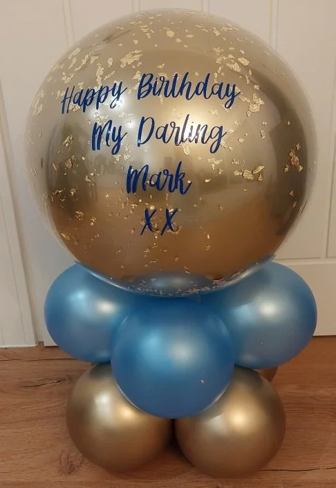 A large transparent balloon with gold confetti and blue lettering that reads "Happy Birthday My Darling Mark xx" sits atop a cluster of smaller blue and gold balloons.