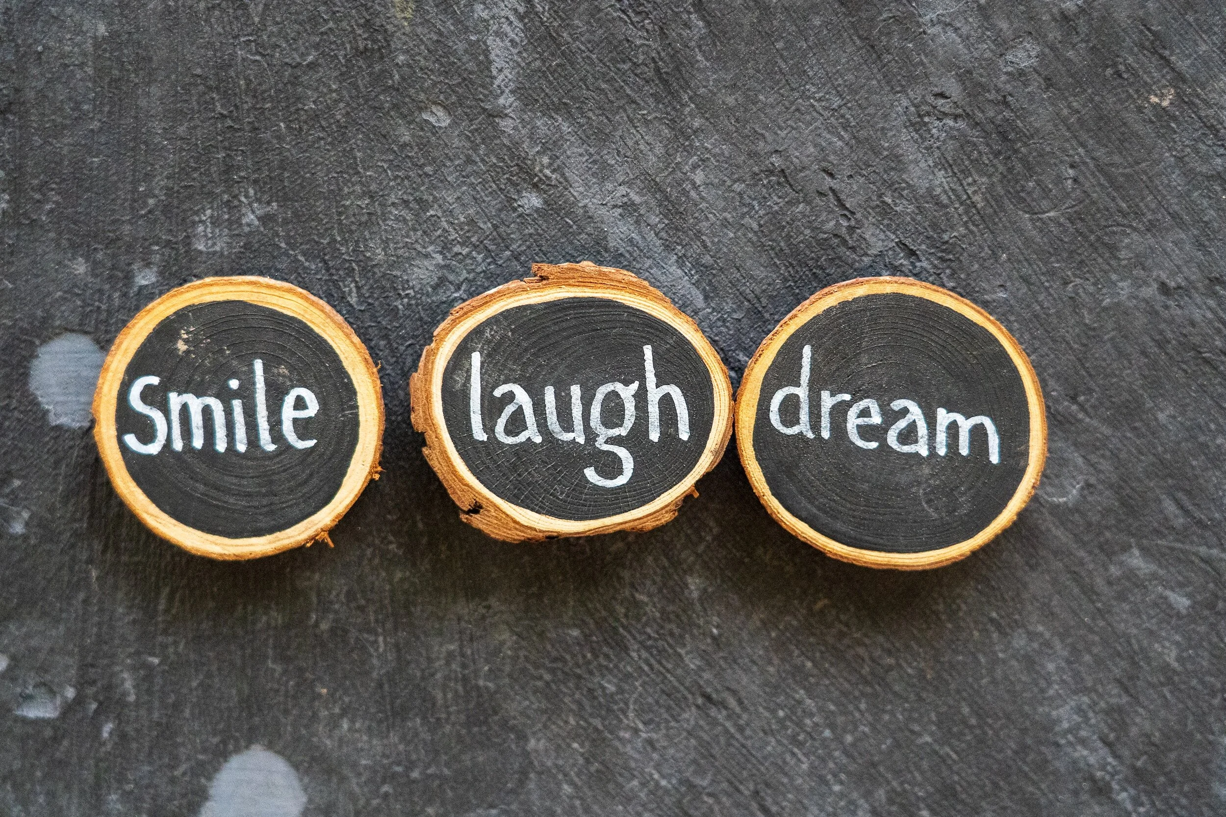 Three wooden slices with black chalkboard backgrounds and white handwritten words: "Smile," "laugh," and "dream," arranged on a dark textured surface.